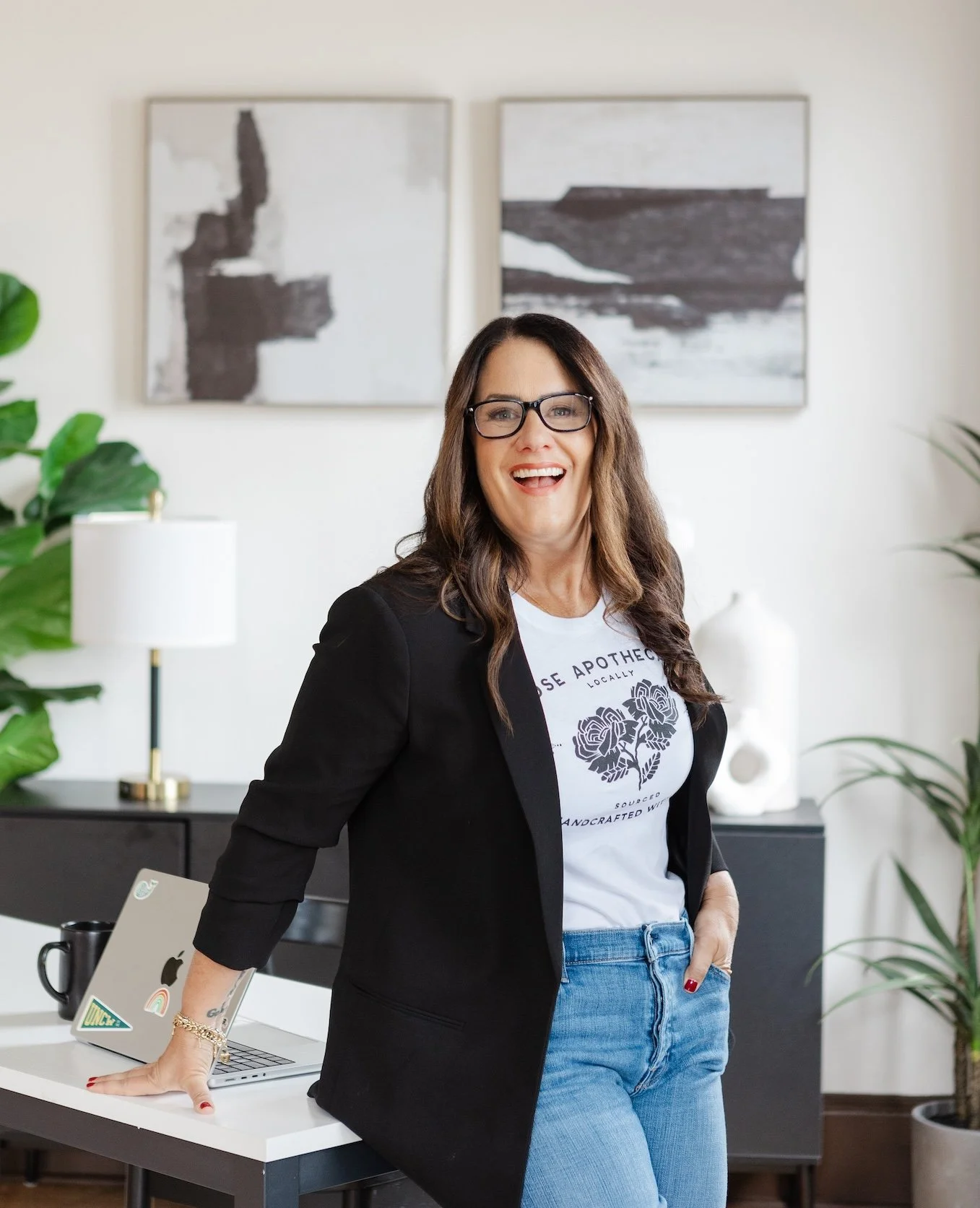 A woman with long brown hair and glasses smiling in a modern office, wearing a black blazer, T-shirt, and jeans, standing behind a desk with a laptop and mug.