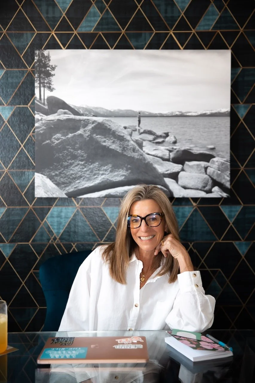 A woman with glasses and blonde hair smiling while sitting at a desk in front of a geometric patterned wall and black and white landscape photograph, with a laptop, a notebook, and a pen on the desk.