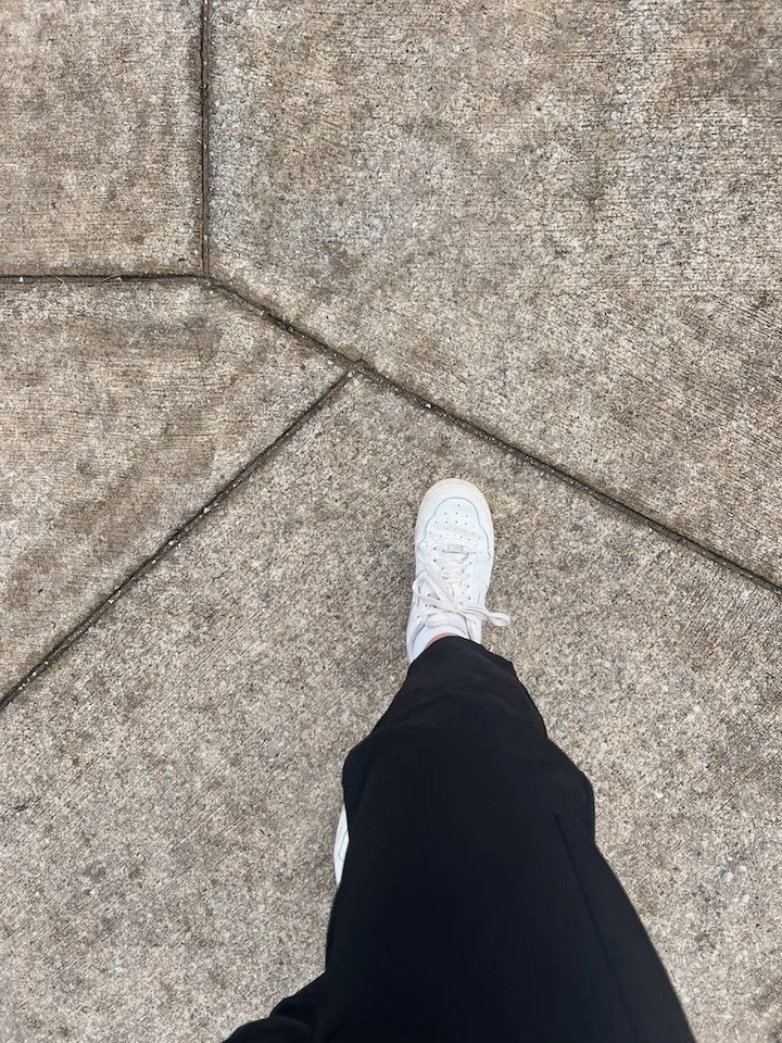 A person's legs and feet in white sneakers and black pants walking on a sidewalk with concrete slabs.