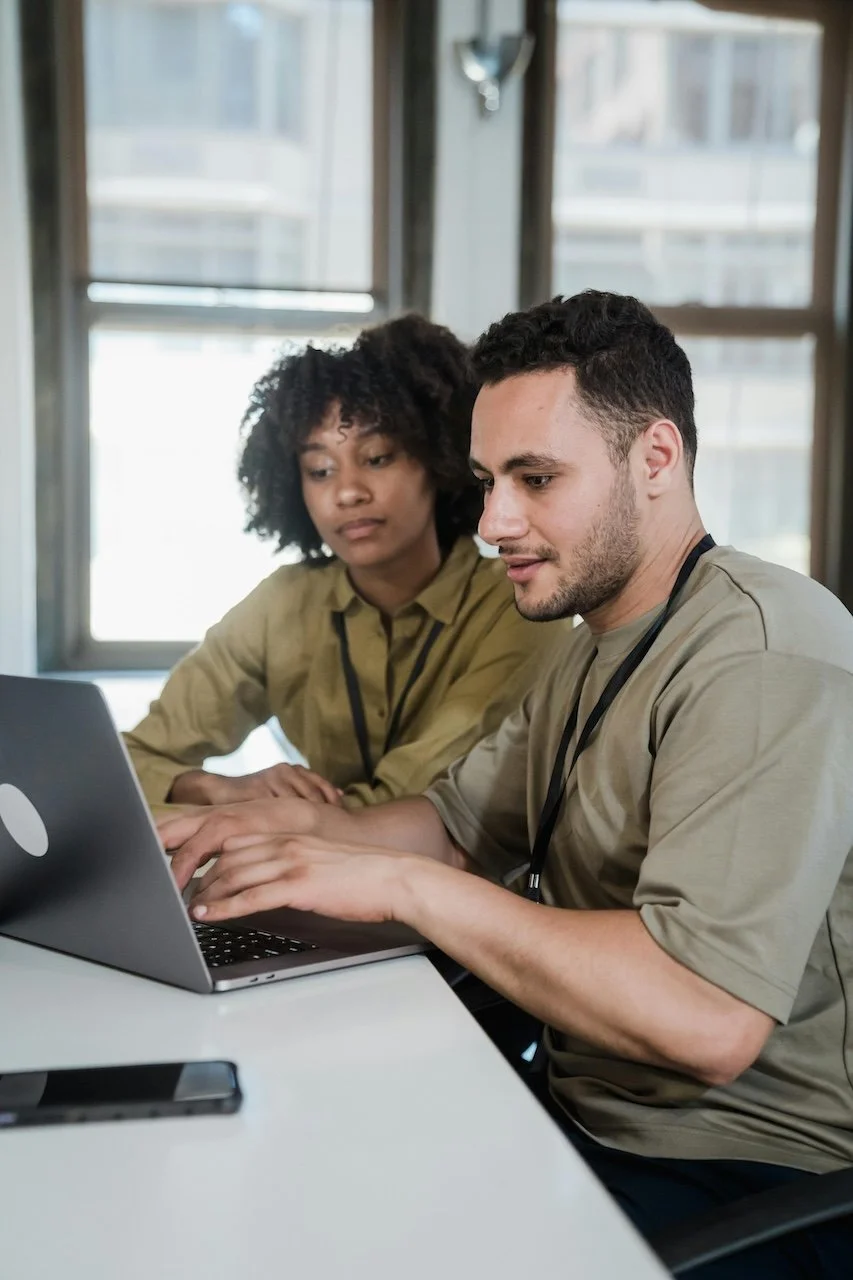 Two people working together on a laptop in an office setting, with large windows in the background.
