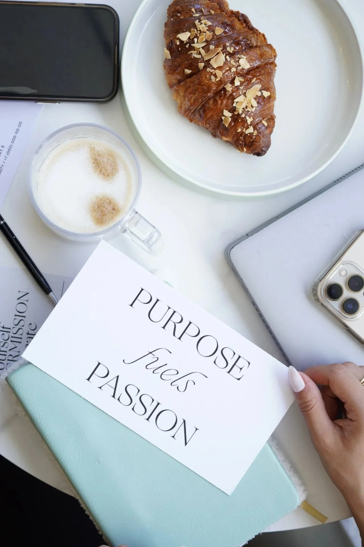 A breakfast scene with a croissant topped with almonds, a cup of frothy coffee, two smartphones, and a handwritten note saying 'Purpose fuels passion,' on a white table.
