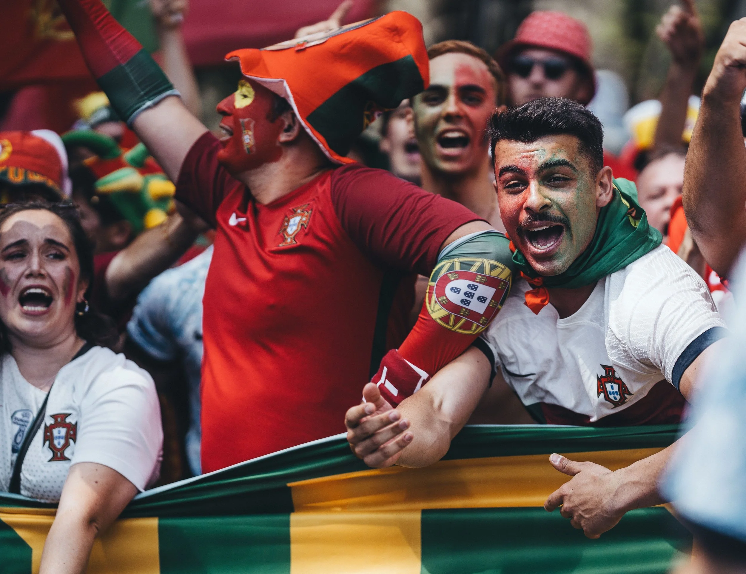 A group of excited soccer fans celebrating, dressed in red, white, and green jerseys and hats, some with painted faces, cheering and waving their arms.