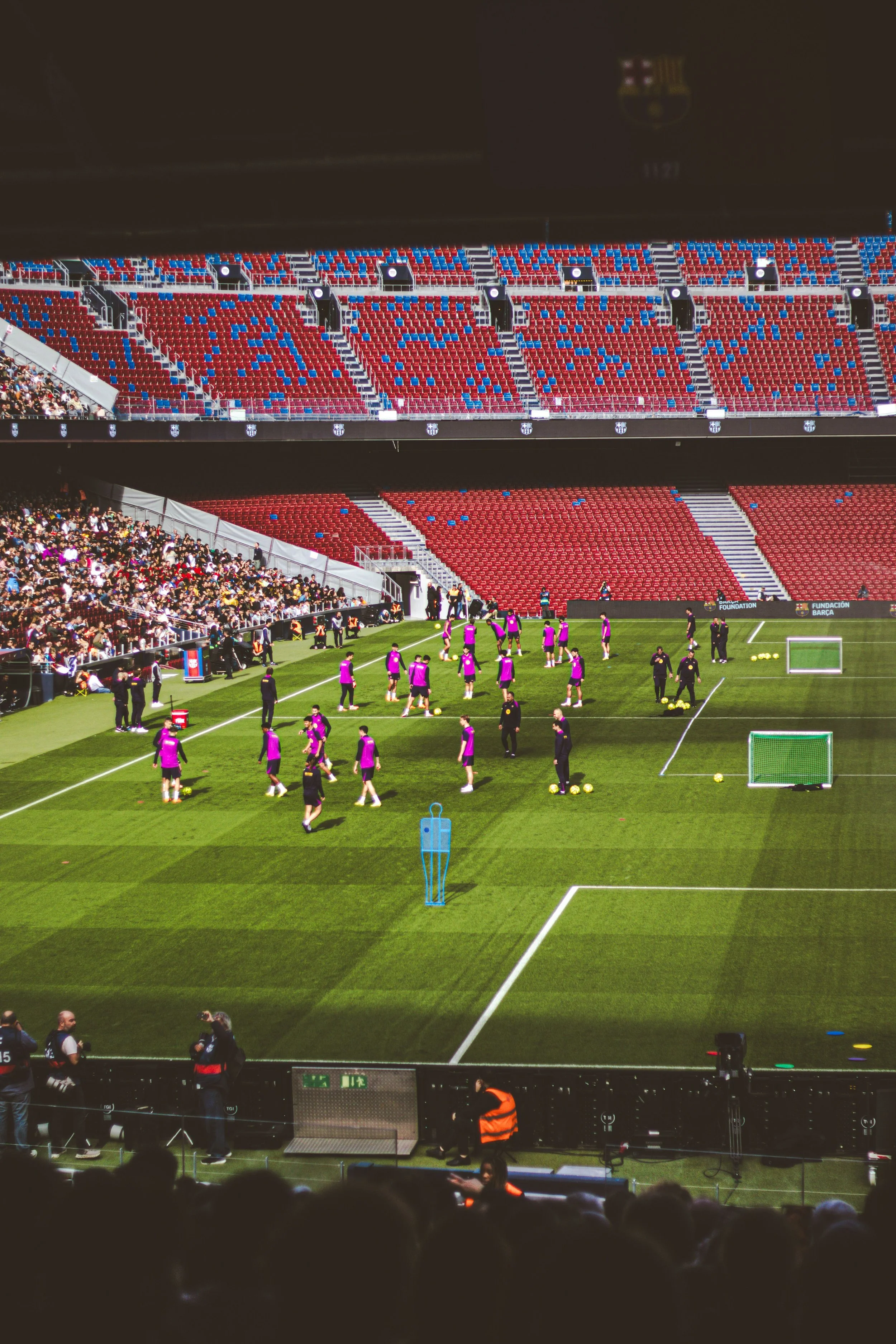 Soccer players warming up on a green field before a match, with spectators in the stands and security personnel nearby.