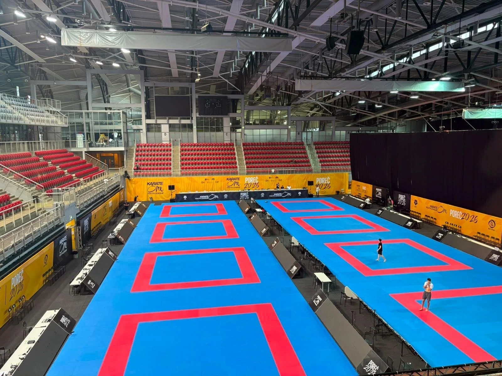 An indoor sports arena set up for karate competitions with multiple blue competition mats with red boundaries, seating areas with red chairs, and yellow banners displaying logos and event information.