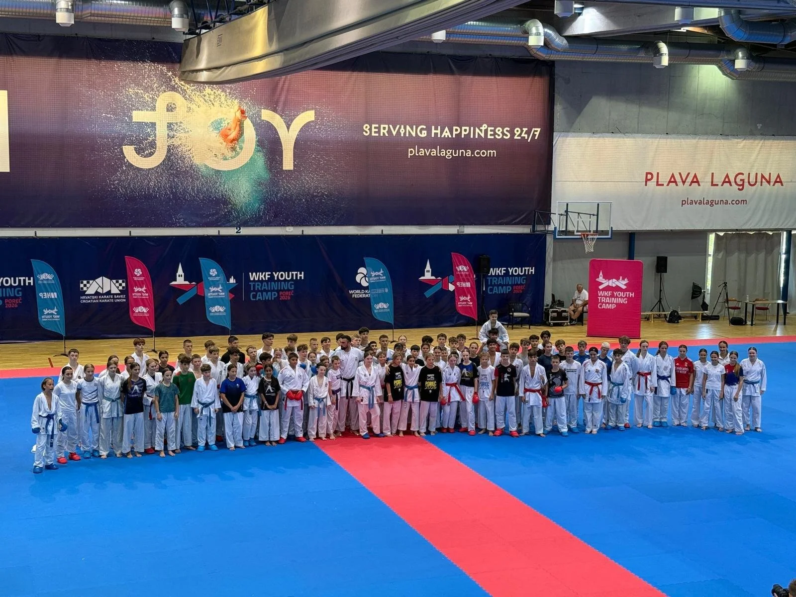 Group of young martial artists in uniform standing on a blue and red mat in an indoor gymnasium, participating in a training camp event.
