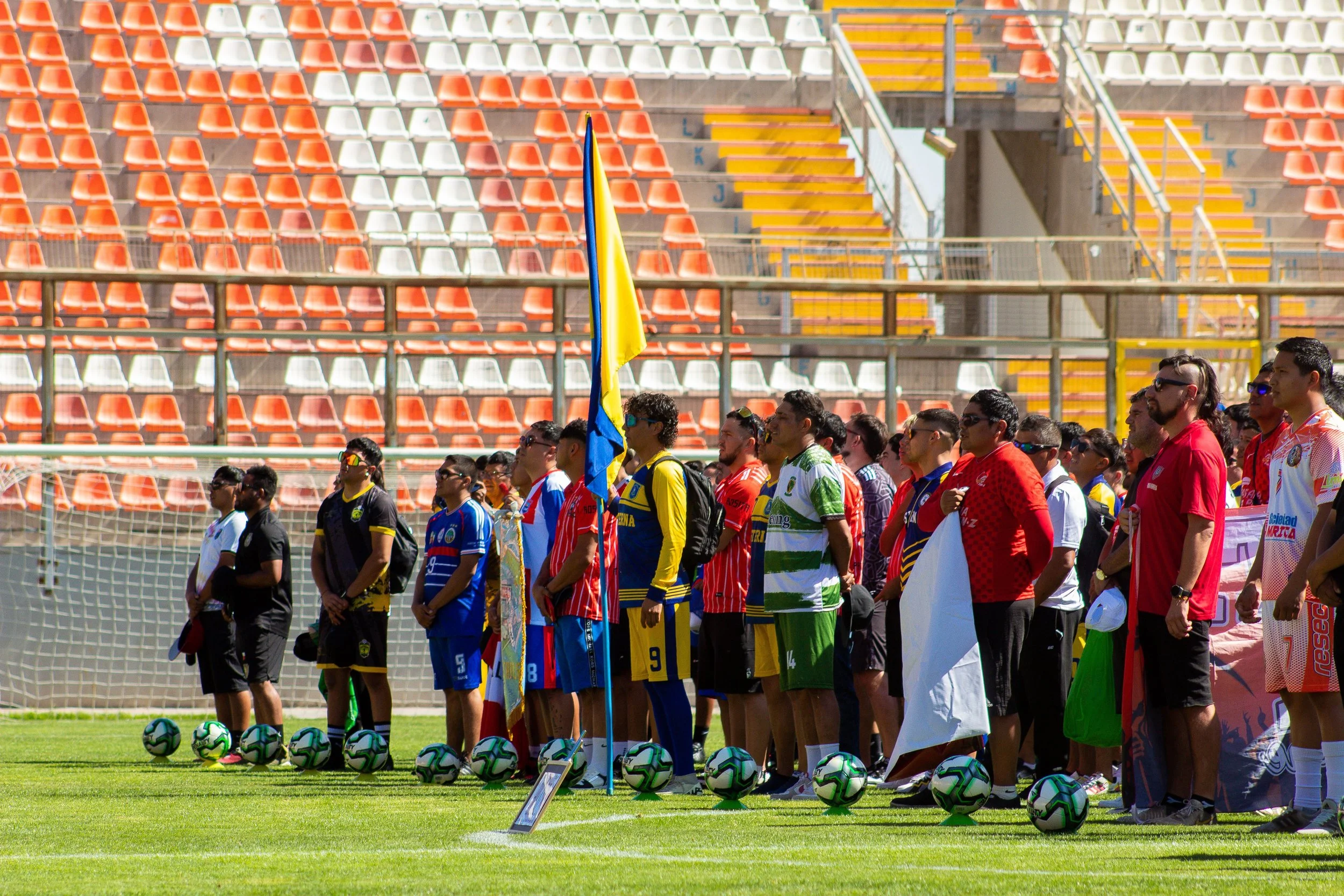 Soccer players dressed in colorful team uniforms stand in a line on a soccer field, holding flags and surrounded by numerous soccer balls, with orange and white stadium seating in the background.