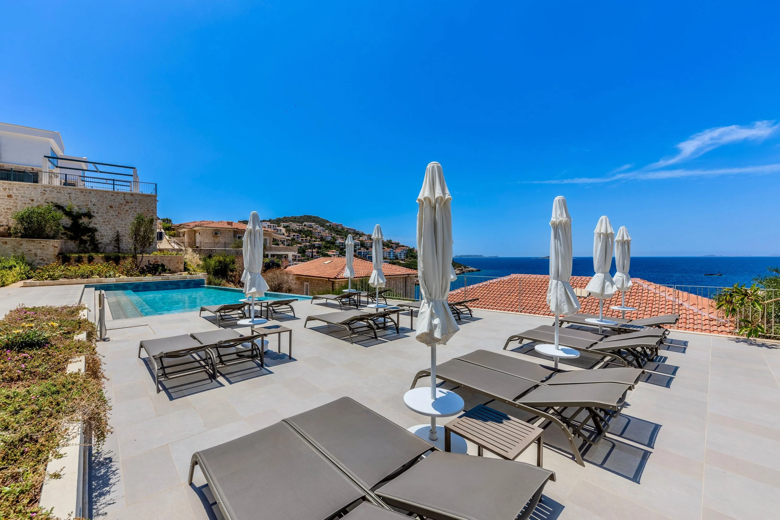 View of a luxurious outdoor pool area with lounge chairs and umbrellas overlooking a hillside and the ocean under a clear blue sky.