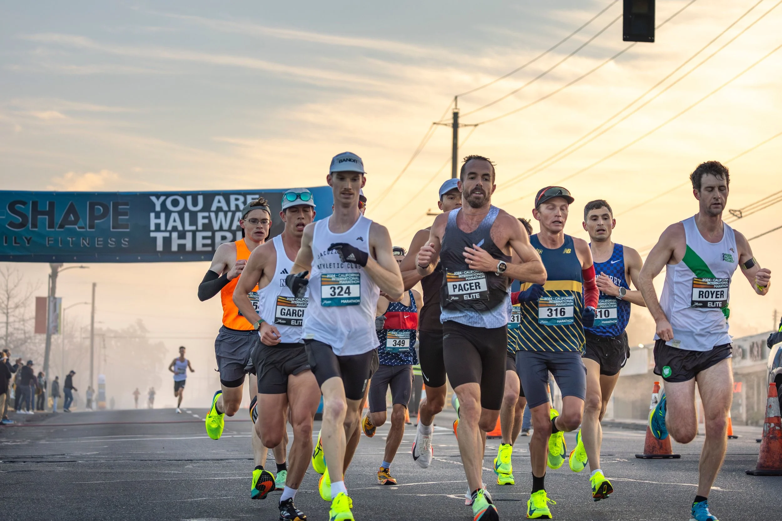 A group of runners participating in a marathon race, running on a city street during sunset. Some runners wear race bibs and athletic gear.