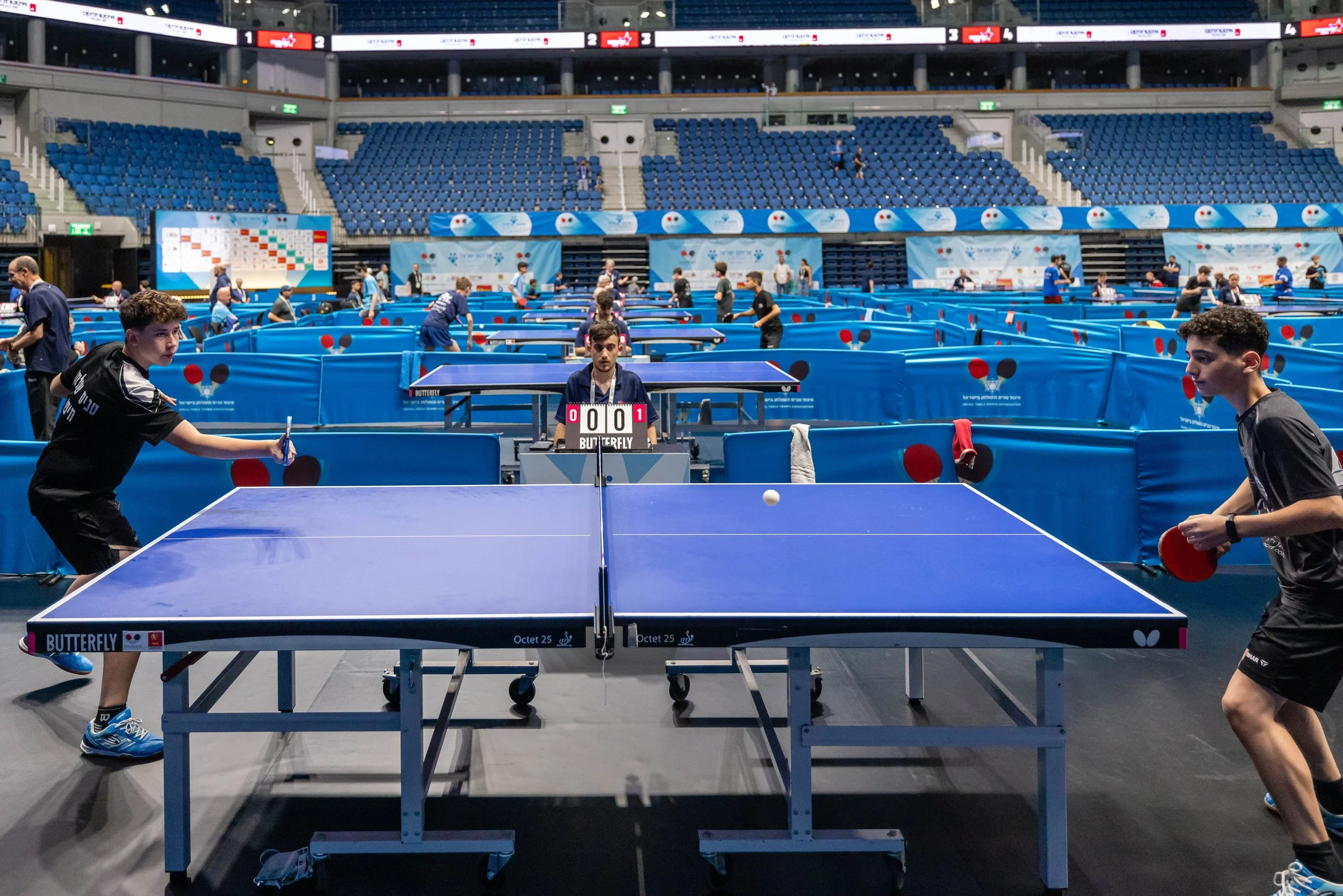 An indoor table tennis arena with multiple tables, blue barriers, and players practicing. Several spectators sit in the stands, and there are multiple digital scoreboards and banners in the background.