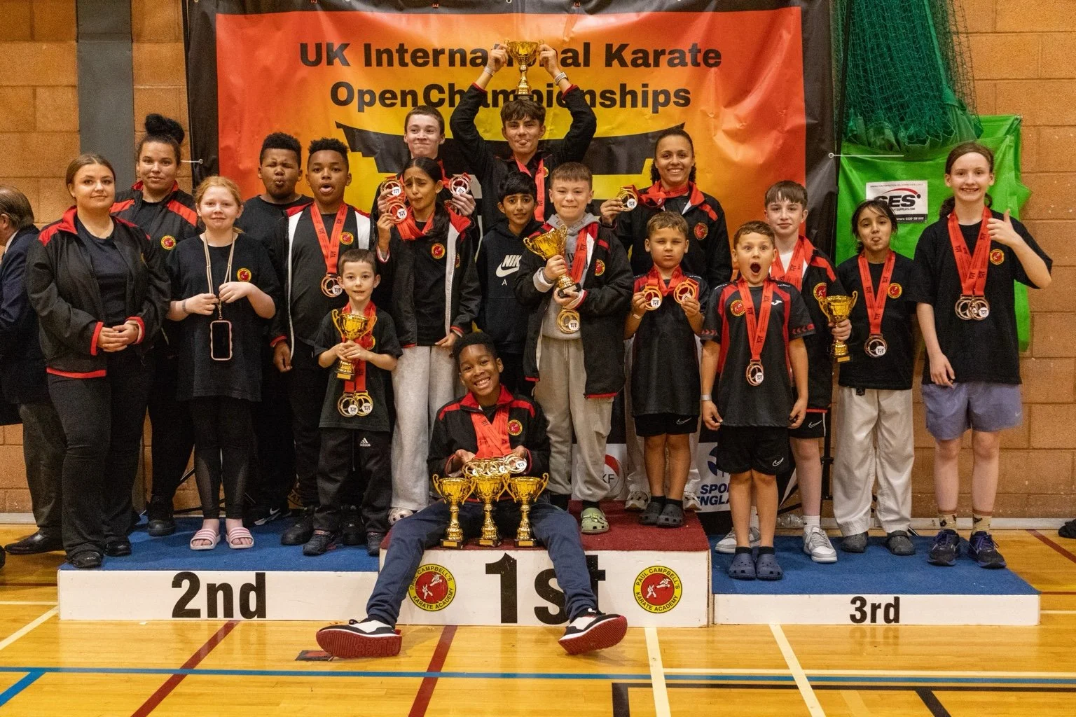 Young karate competitors and instructors celebrating on the winners' podium at the UK International Karate Open Championships, with medals and trophies, inside a gymnasium.