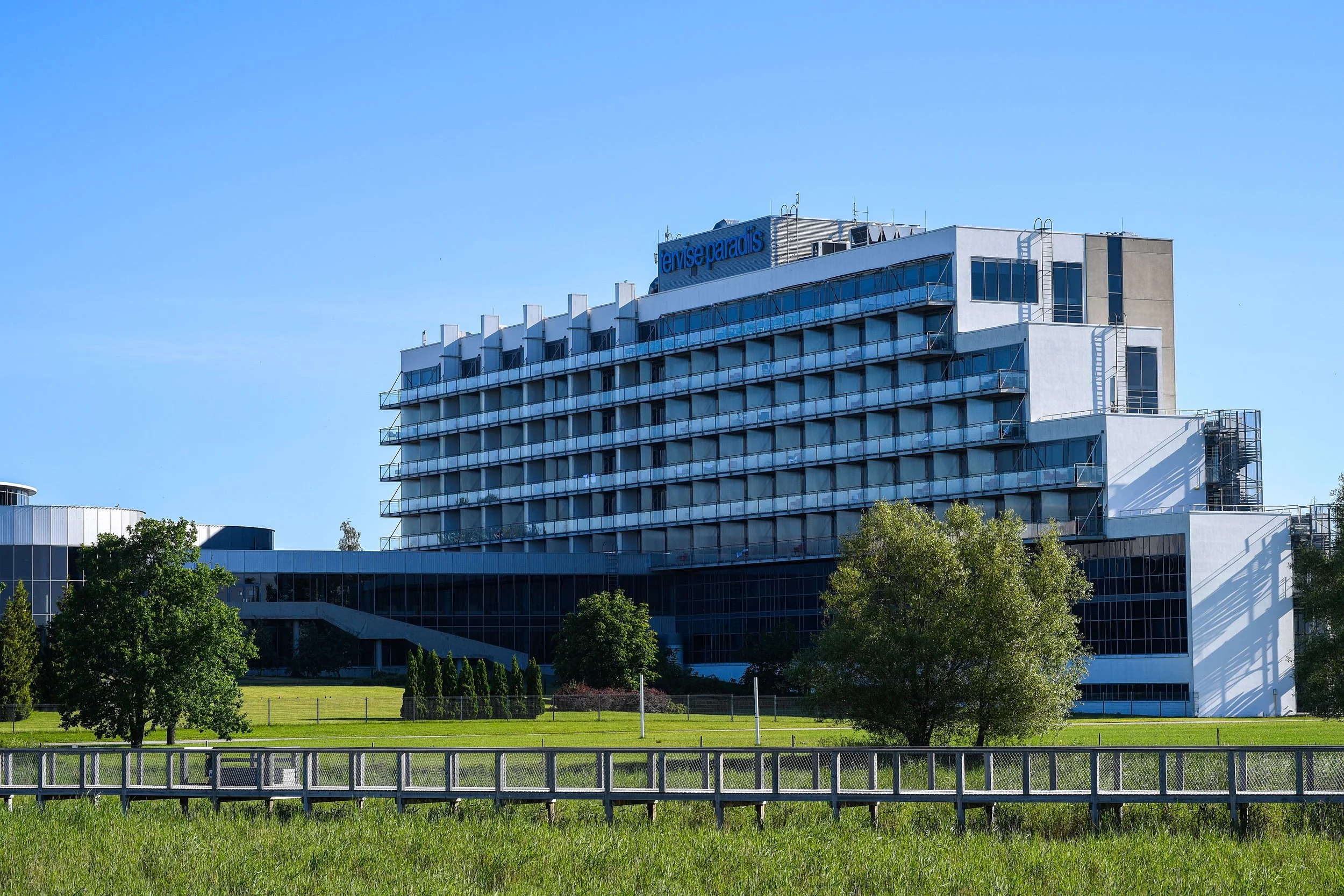 Modern multi-story building with balconies and glass windows, surrounded by greenery and trees, under a clear blue sky.