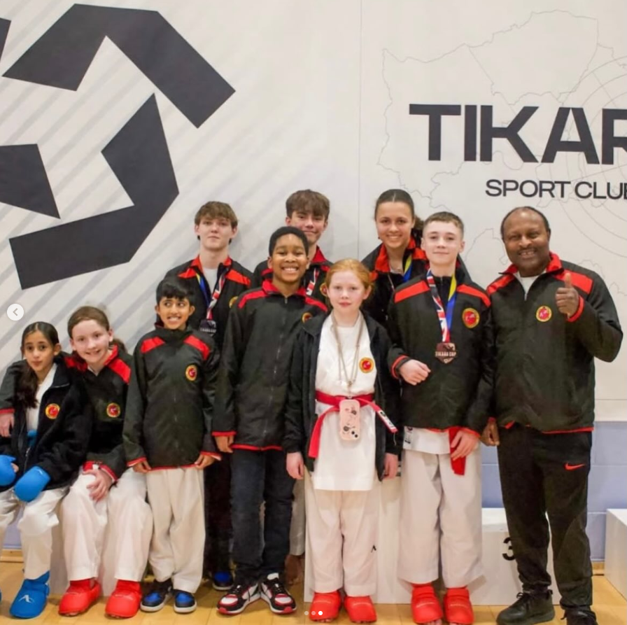 A group of children and an adult coach posing together at Tikar Sport Club, some with medals and martial arts uniforms, standing inside the club with a logo and map background.