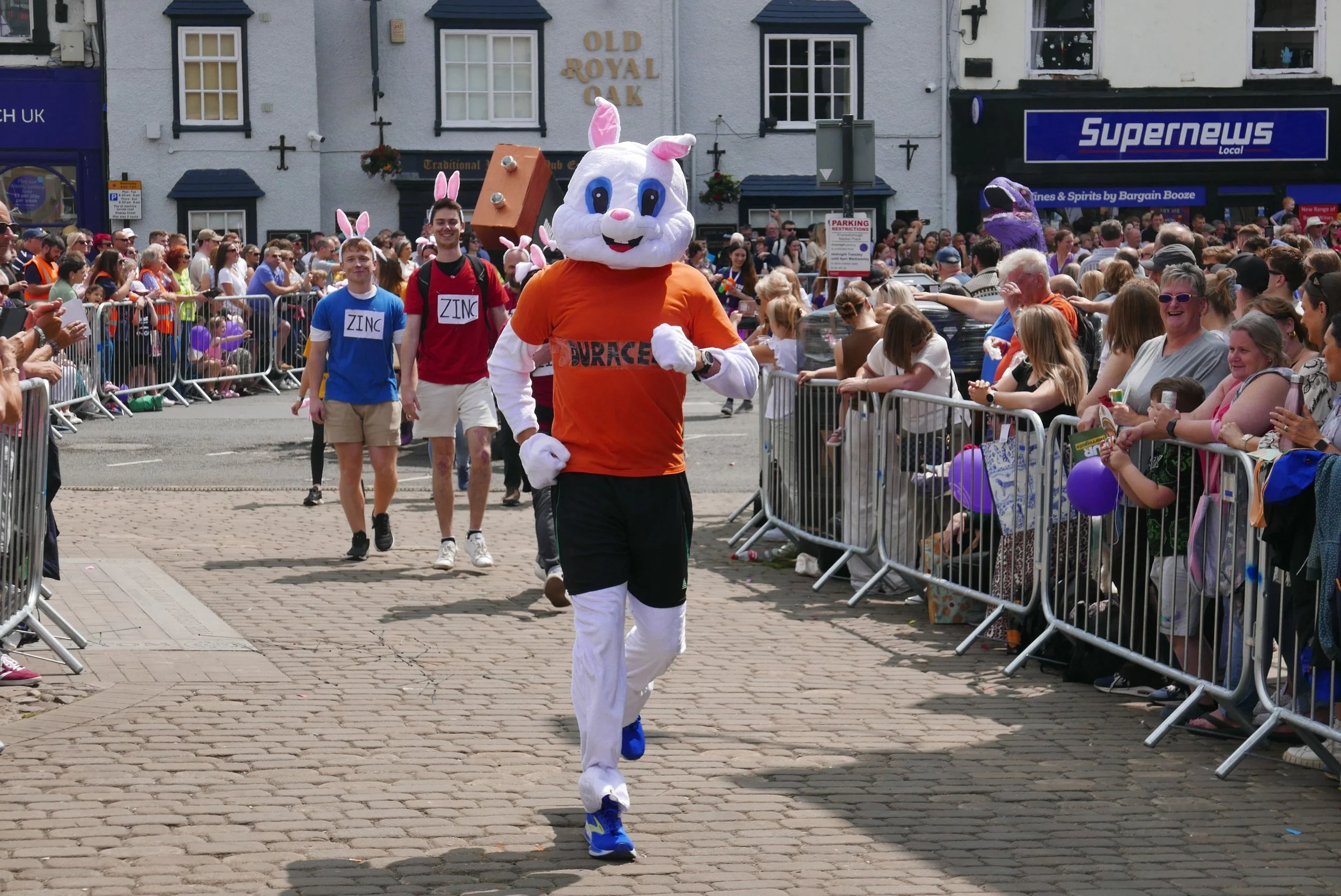 Person in a bunny costume running in a parade, with a crowd of spectators behind barriers watching and cheering.