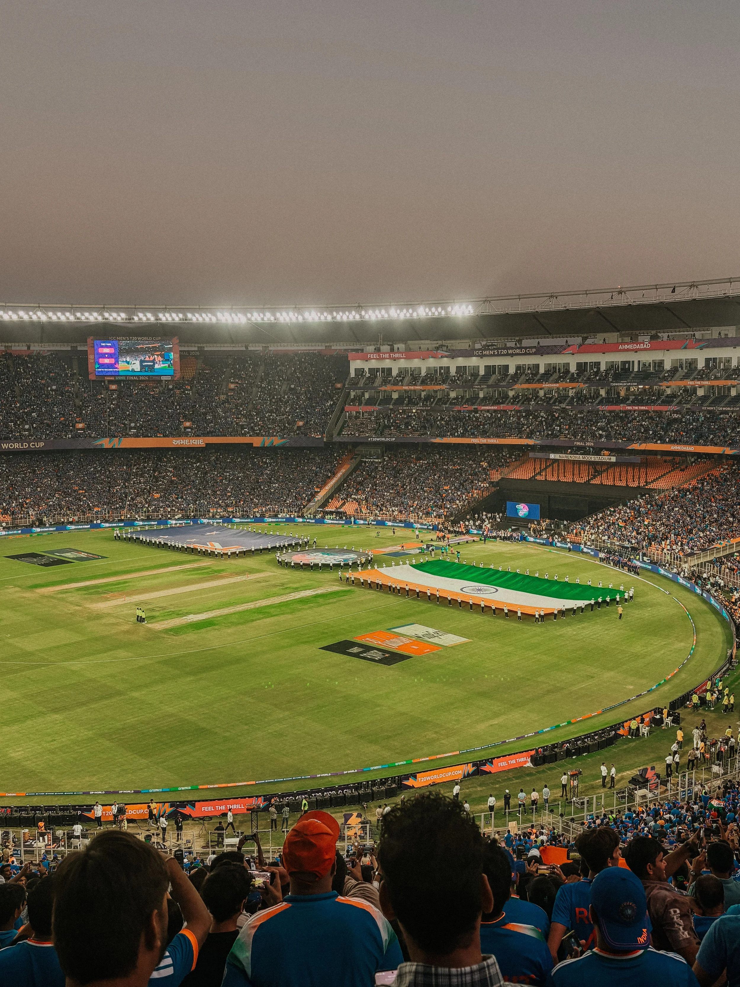 Crowd of spectators in a stadium watching a cricket match, with the Indian flag displayed on the field and banners around the stadium, during the ICC Men's T20 World Cup in Ahmedabad.