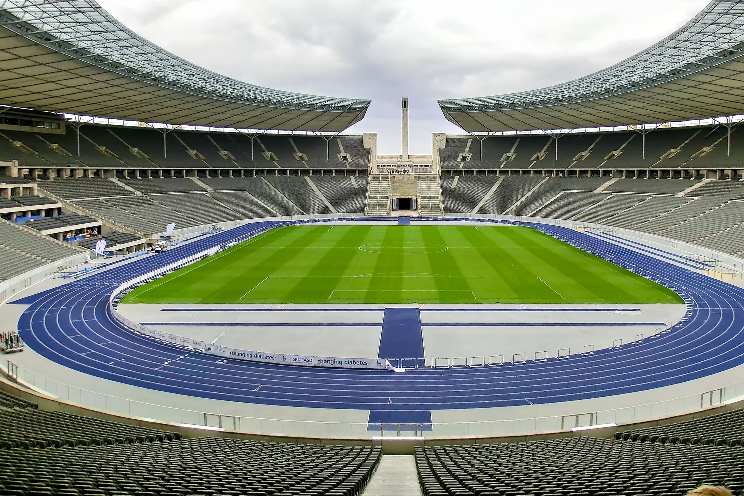 Empty sports stadium with a green field and a blue running track, covered stands, and a cloudy sky overhead.