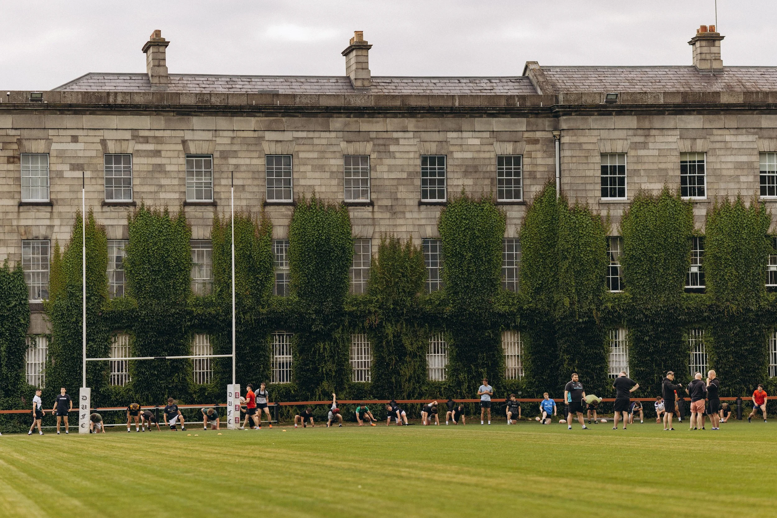 People playing rugby on a field with a large stone building with windows and ivy in the background.