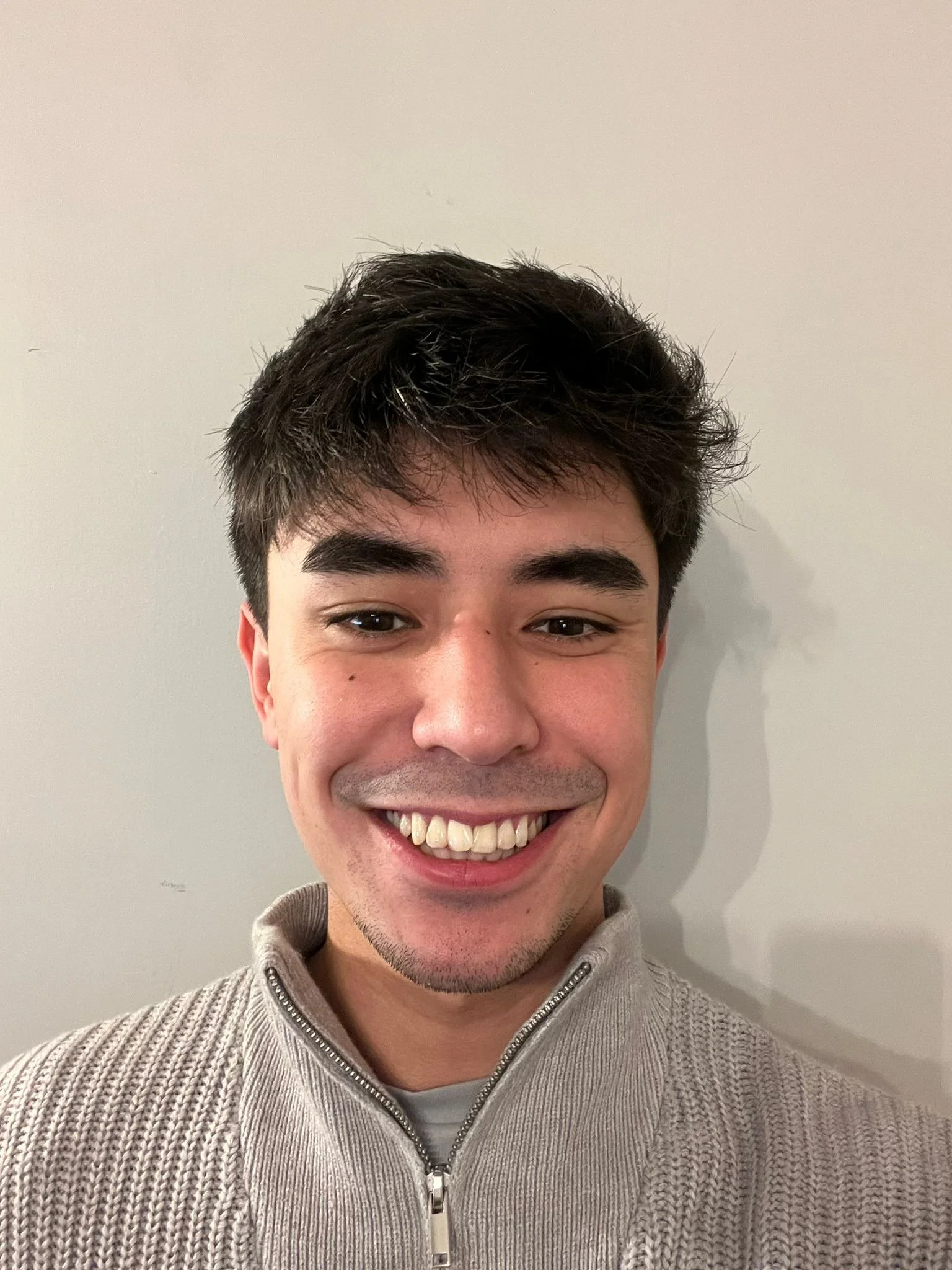 A smiling young man with dark hair and eyebrows, wearing a light gray zip-up sweater against a plain light-colored wall.