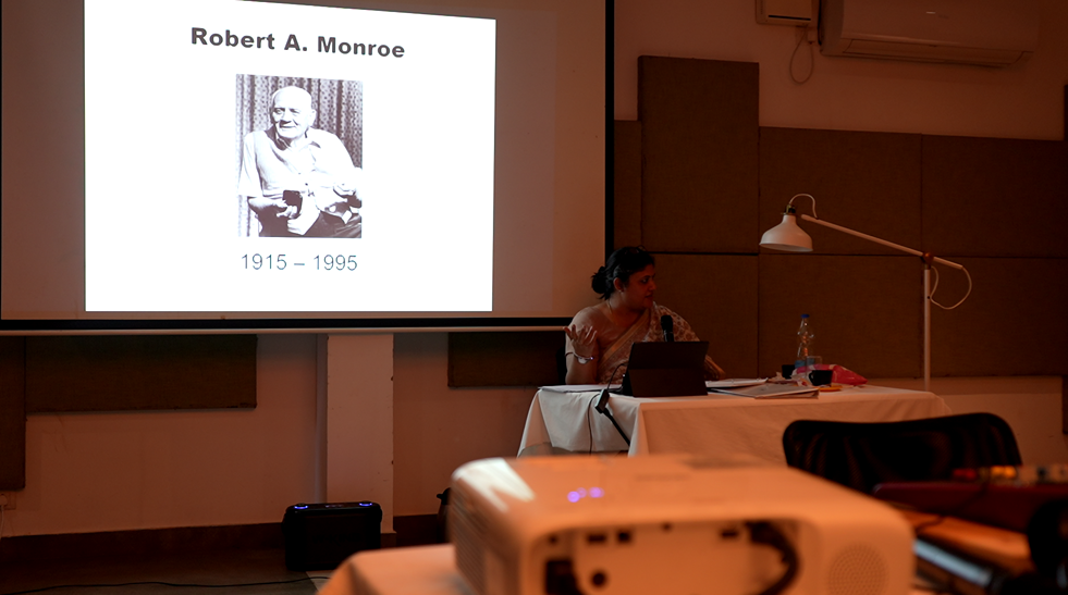 Presentation slide showing a black and white photograph of Robert A. Monroe, with his name and birth and death years (1915-1995). Dr Rashhi Sharma  addressing students of Monroe training.
