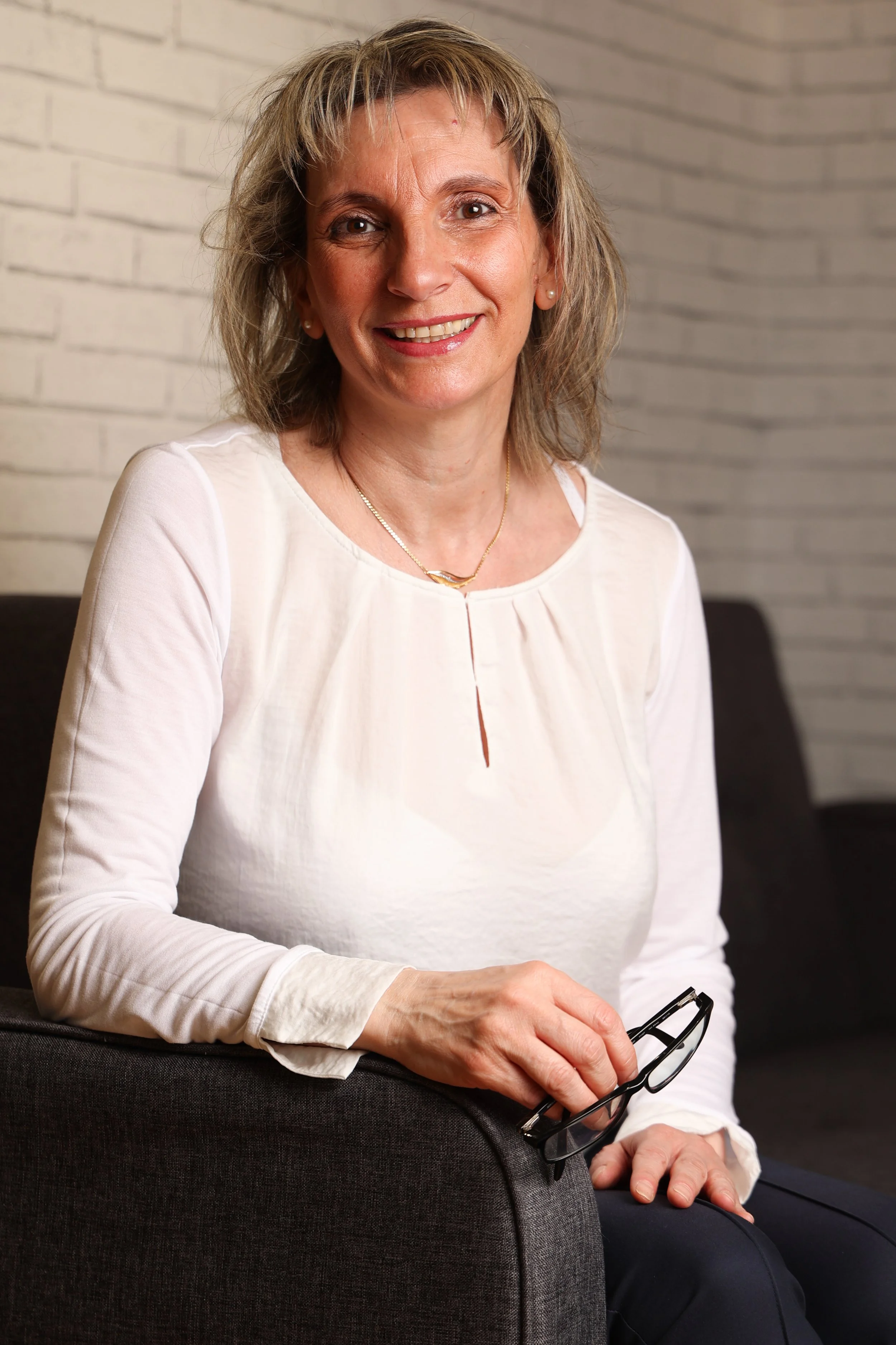 Claudia Hahn-Sauren sitzt mit einer Brille in der Hand auf einem Sofa und lächelt freundlich. Als Heilpraktikerin für Psychotherapie in Mainz bilden fundierte Ausbildungen und regelmäßige Fortbildungen die Grundlage ihrer Arbeit.