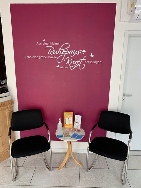 Wartebereich der Sanadios Ganzheitliche Naturheilpraxis in Mainz-Hechtsheim mit zwei schwarzen Sesseln vor einer farbigen Wand mit dem Schriftzug „Aus einer kleinen Ruhepause kann eine große Quelle neuer Kraft entspringen“.