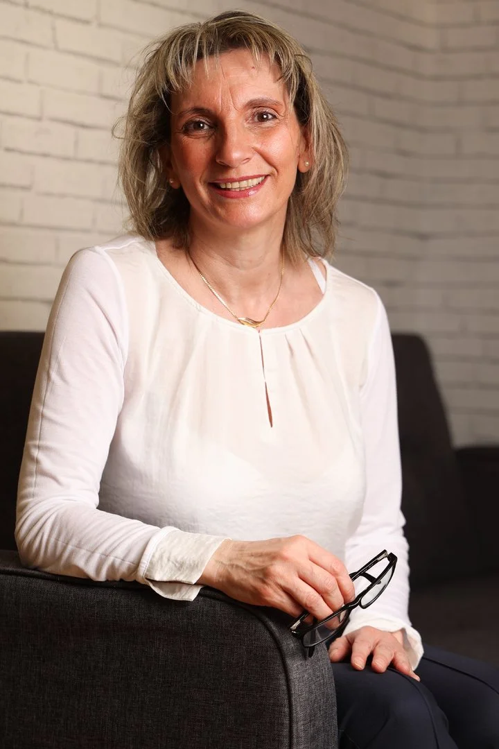 Claudia Hahn-Sauren sitzt mit einer Brille in der Hand auf einem Sofa und lächelt freundlich. Heilpraktikerin für Psychotherapie und Coach in Mainz.