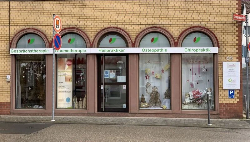 Außenansicht der Sanadios Ganzheitliche Naturheilpraxis in Mainz-Hechtsheim mit Eingang, bodentiefen dekorierten Fenstern und Praxisbeschriftungen für Gesprächstherapie, Traumatherapie, Heilpraktiker, Osteopathie und Chiropraktik.