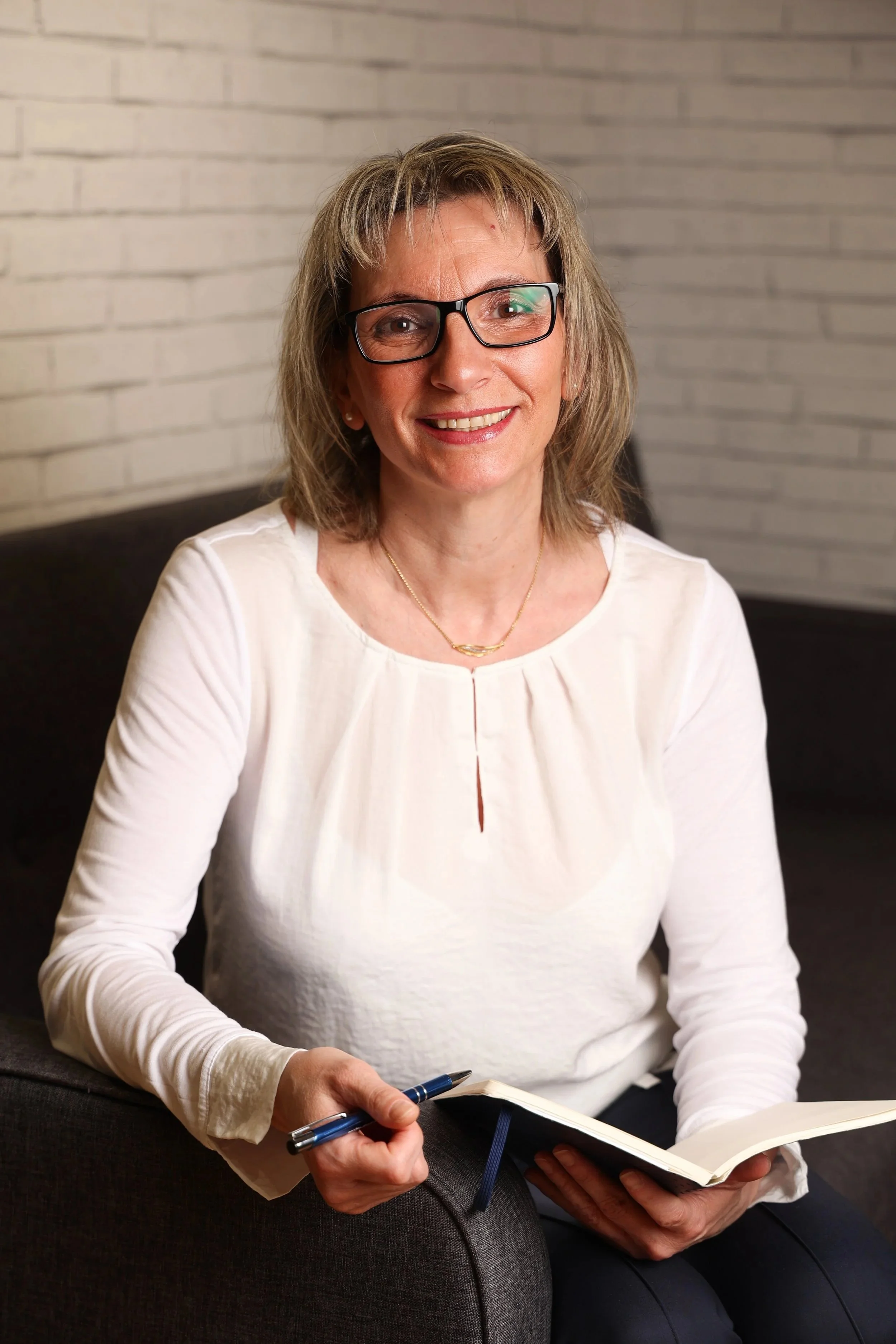 Claudia Hahn-Sauren lächelt freundlich in die Kamera und hält ein Notizbuch und einen Stift in den Händen. Heilpraktikerin für Psychotherapie und Mentalcoach in Mainz.