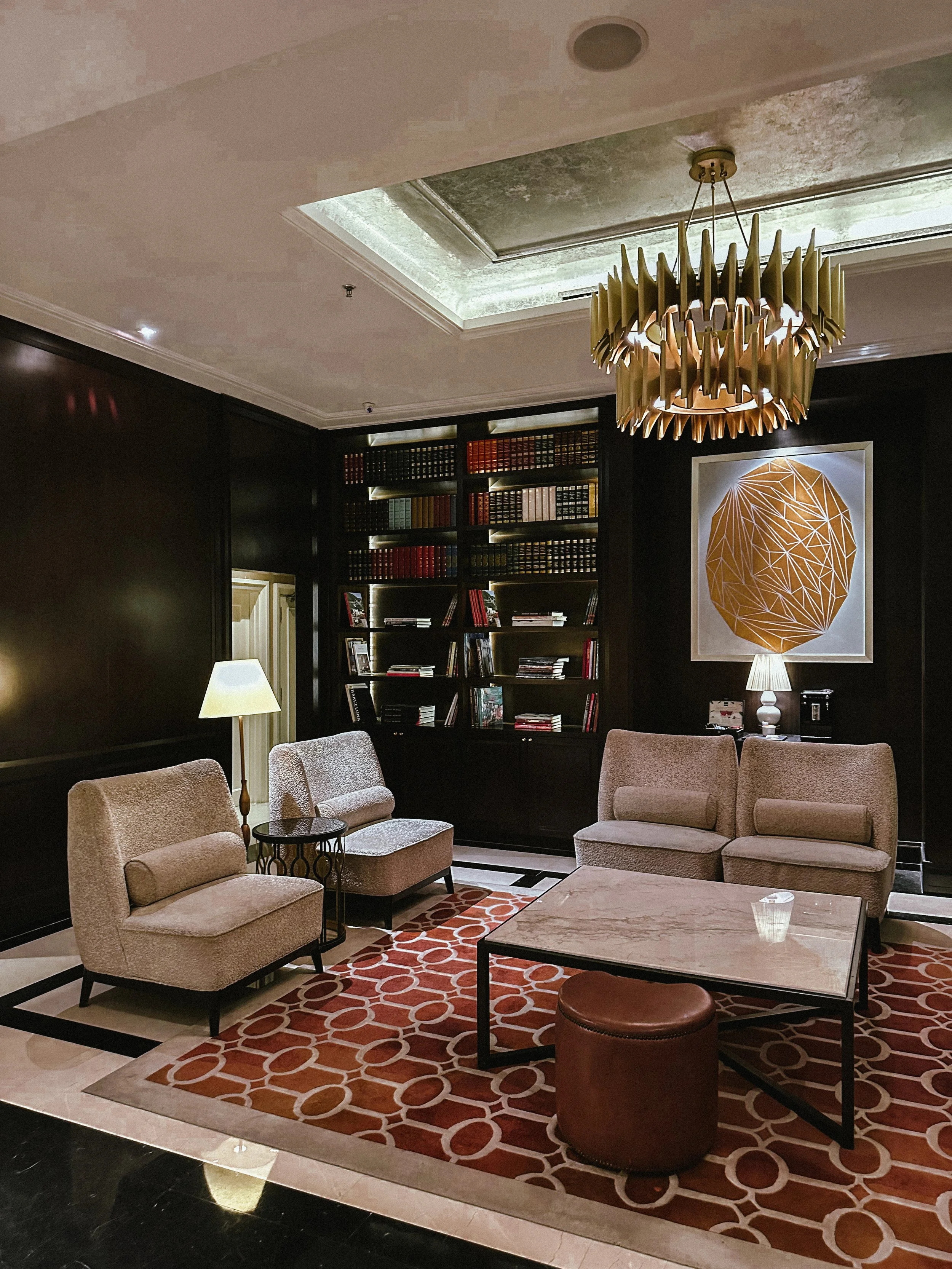 Luxurious hotel lobby with beige seating, modern art on dark wall, bookshelf, marble coffee table, patterned carpet, and elegant lighting fixtures.