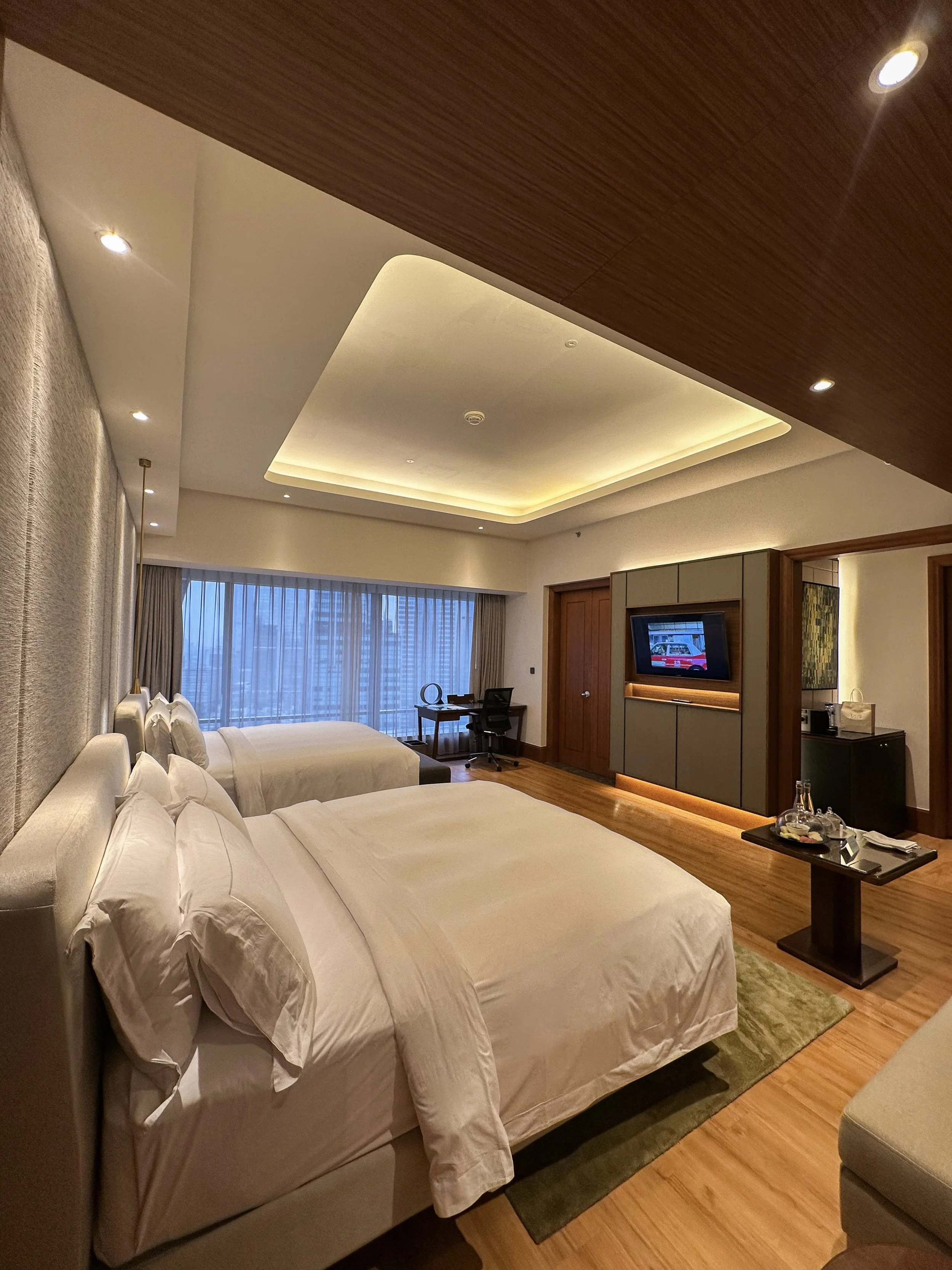 A modern hotel room with two white beds, a large window with curtains, a desk with a chair, a TV on a wall panel, a small table with water bottles and glasses, and warm lighting.