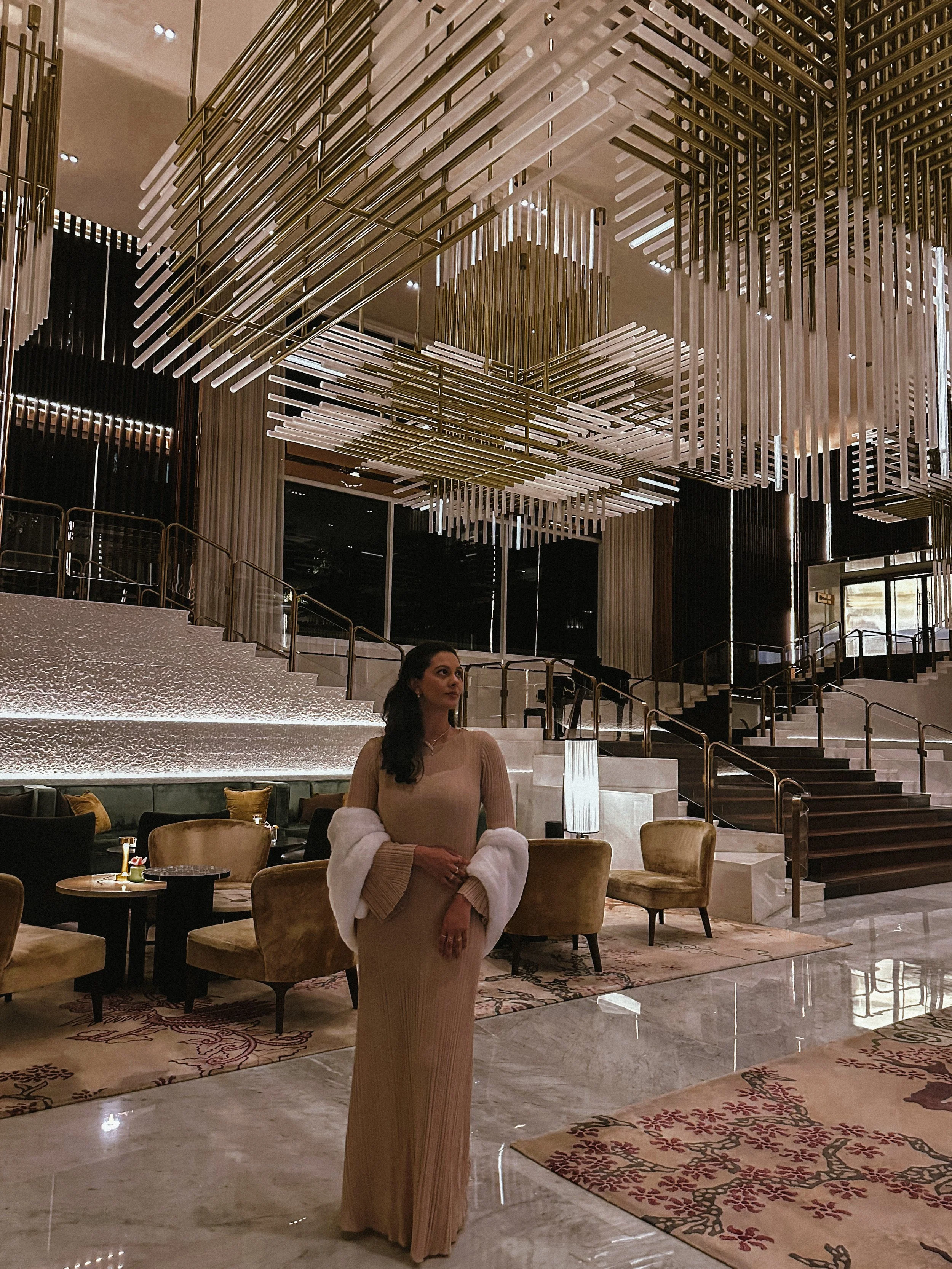 A woman in a beige dress and white fur stole standing in a luxurious hotel lobby with modern chandeliers and elegant seating.