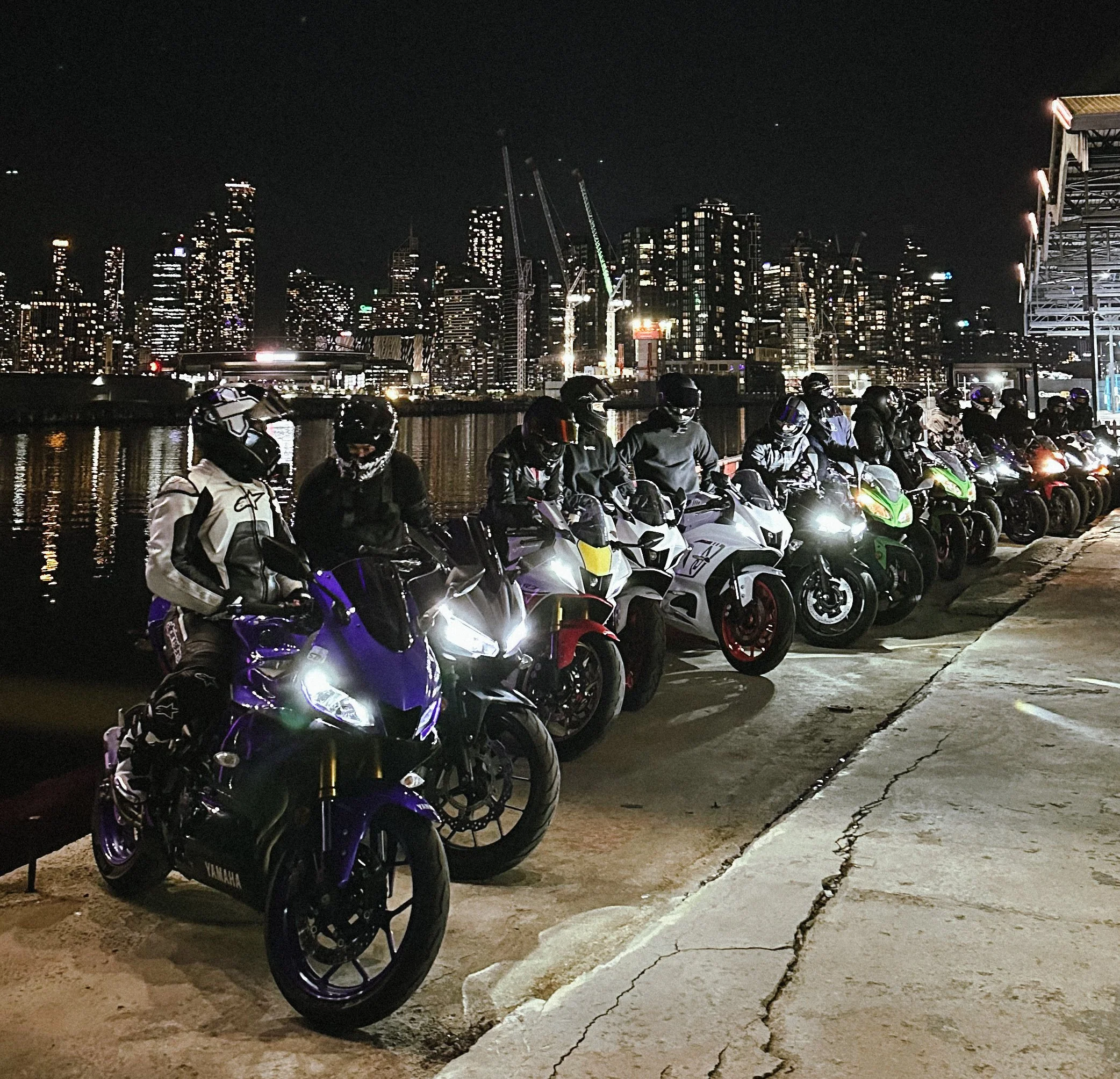 Line of motorbikes parked along a waterfront at night with a city skyline and high-rise buildings in the background.