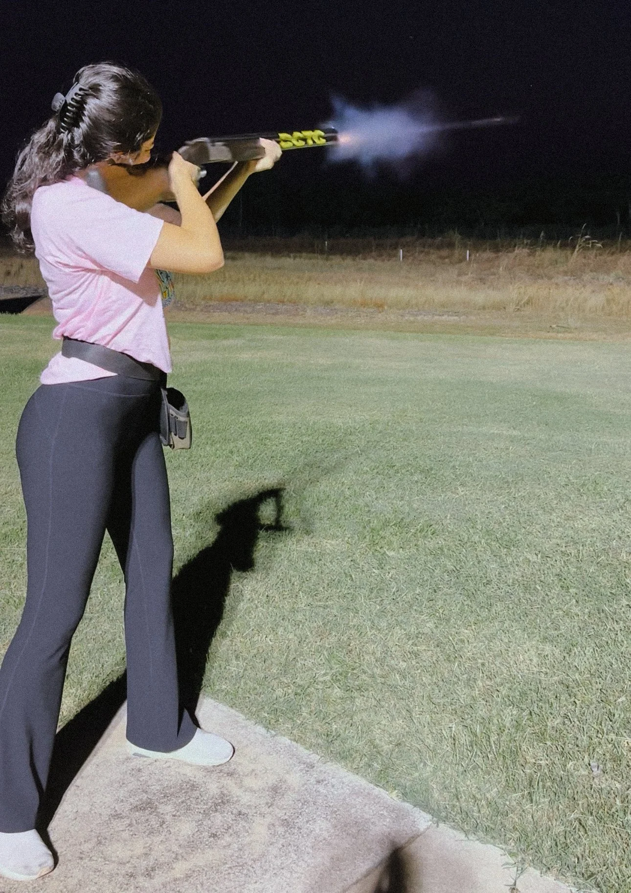 A woman standing on a concrete pad at night, firing a shotgun with smoke and sparks coming from the barrel, on a grassy field.