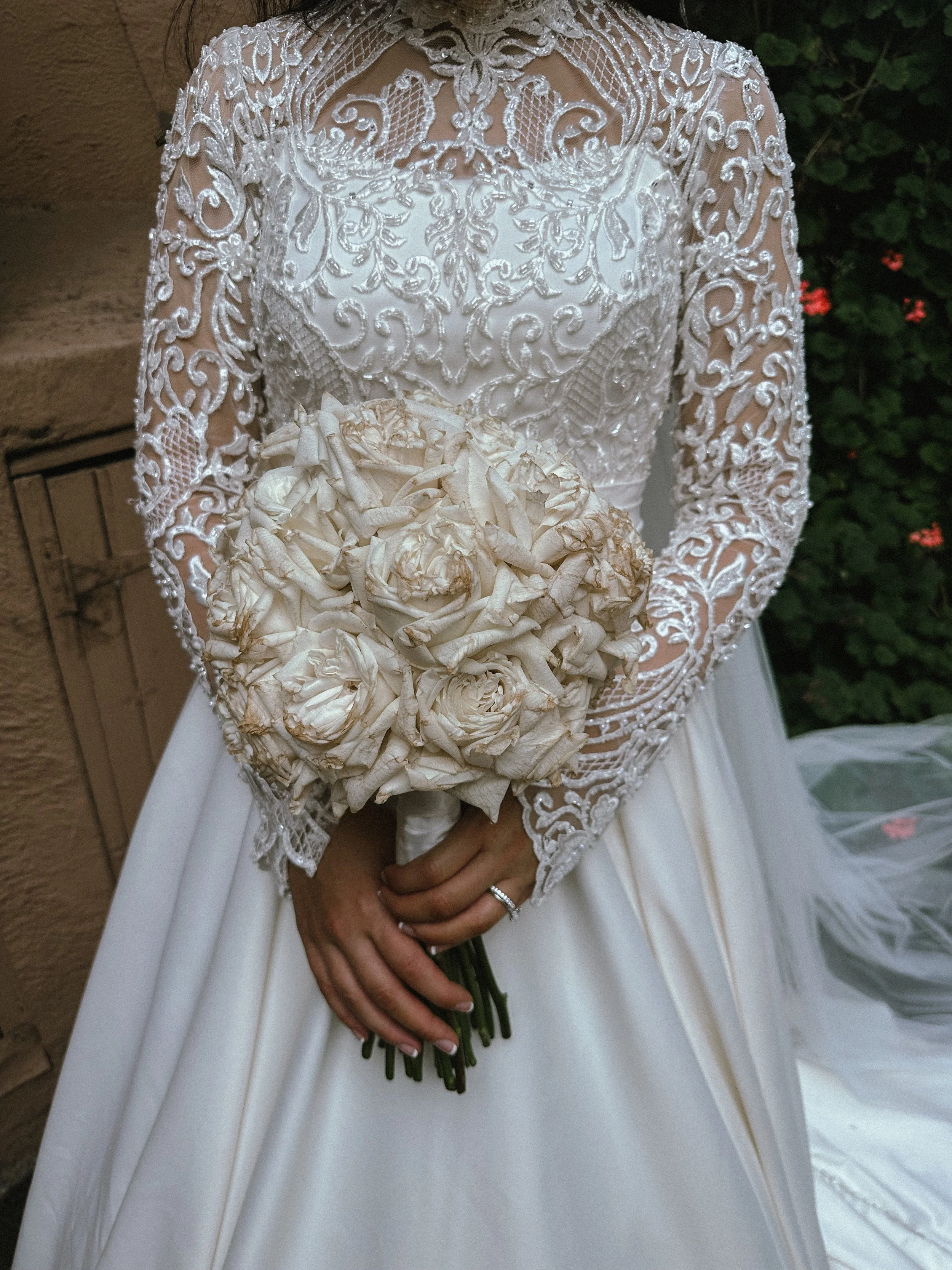 Bride holding a bouquet of wilted white roses, wearing a lace wedding dress with intricate embroidery.