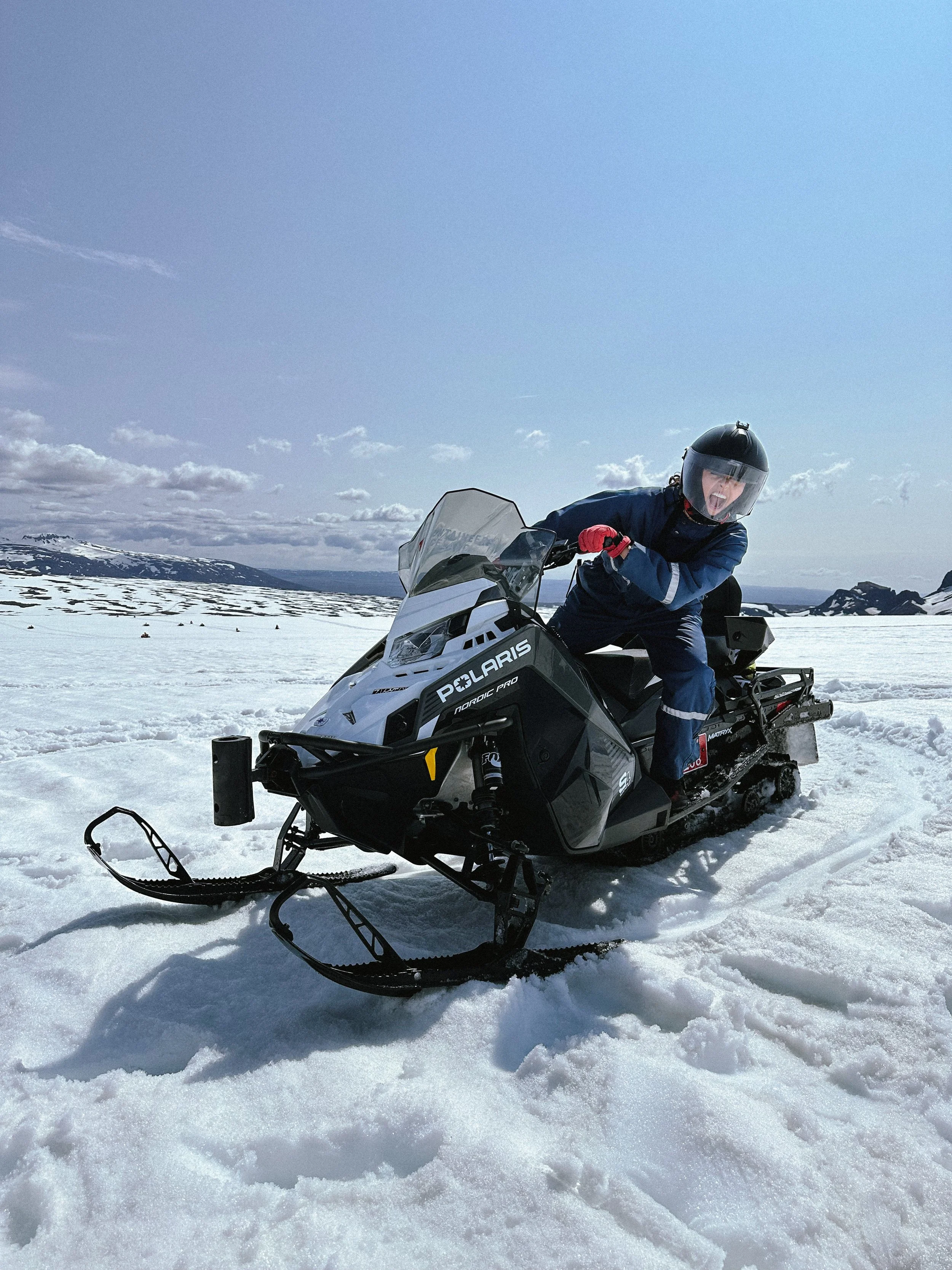 Person wearing helmet and blue clothing riding a Polaris snowmobile on snow-covered landscape with mountains in the background under cloudy sky.