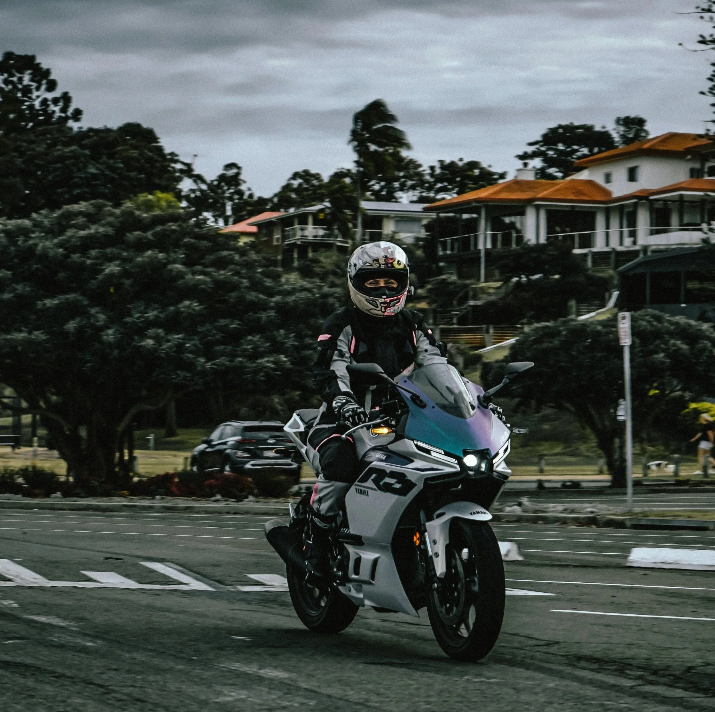 A motorcyclist riding a Yamaha motorcycle on a city street, wearing a helmet and riding gear, with houses and trees in the background on a cloudy day.