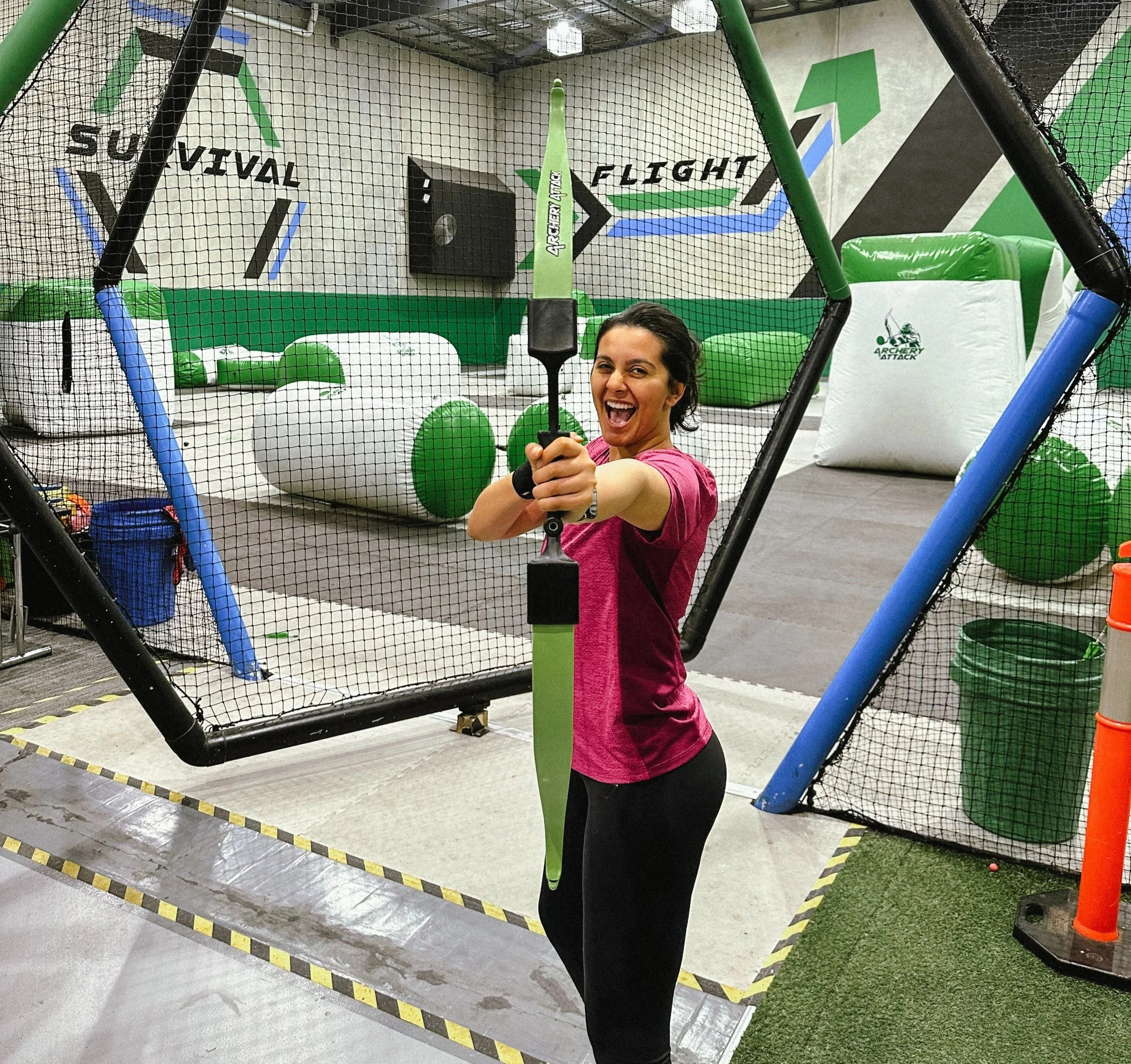 Woman in pink athletic shirt smiling and holding a large foam sword at an indoor axe-throwing or archery range with padded barriers and safety netting.