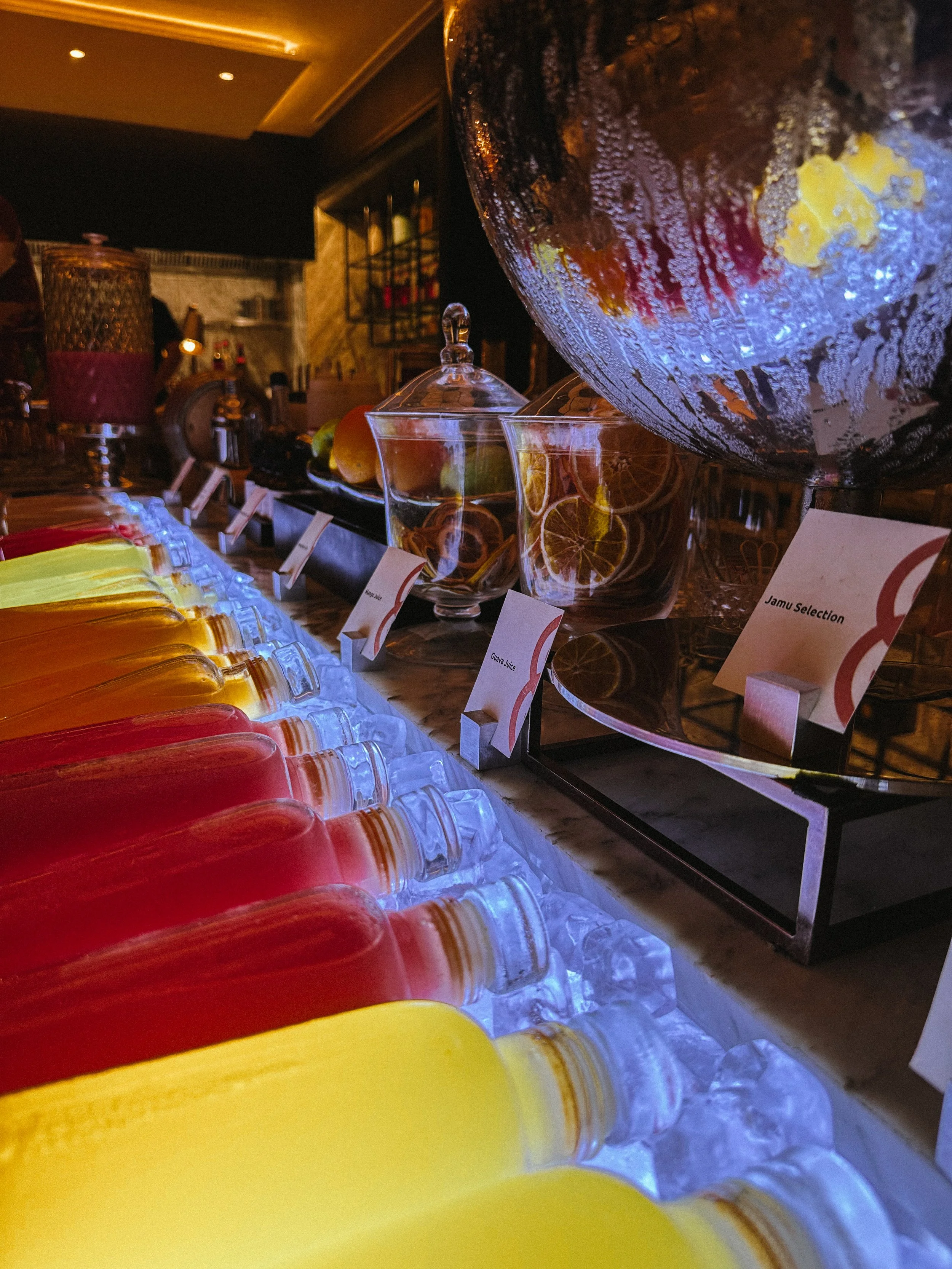 A colorful display of bottled beverages arranged on a table with labels, illuminated by blue lighting underneath, with glass jars and a large glass bowl filled with fruit slices in the background.