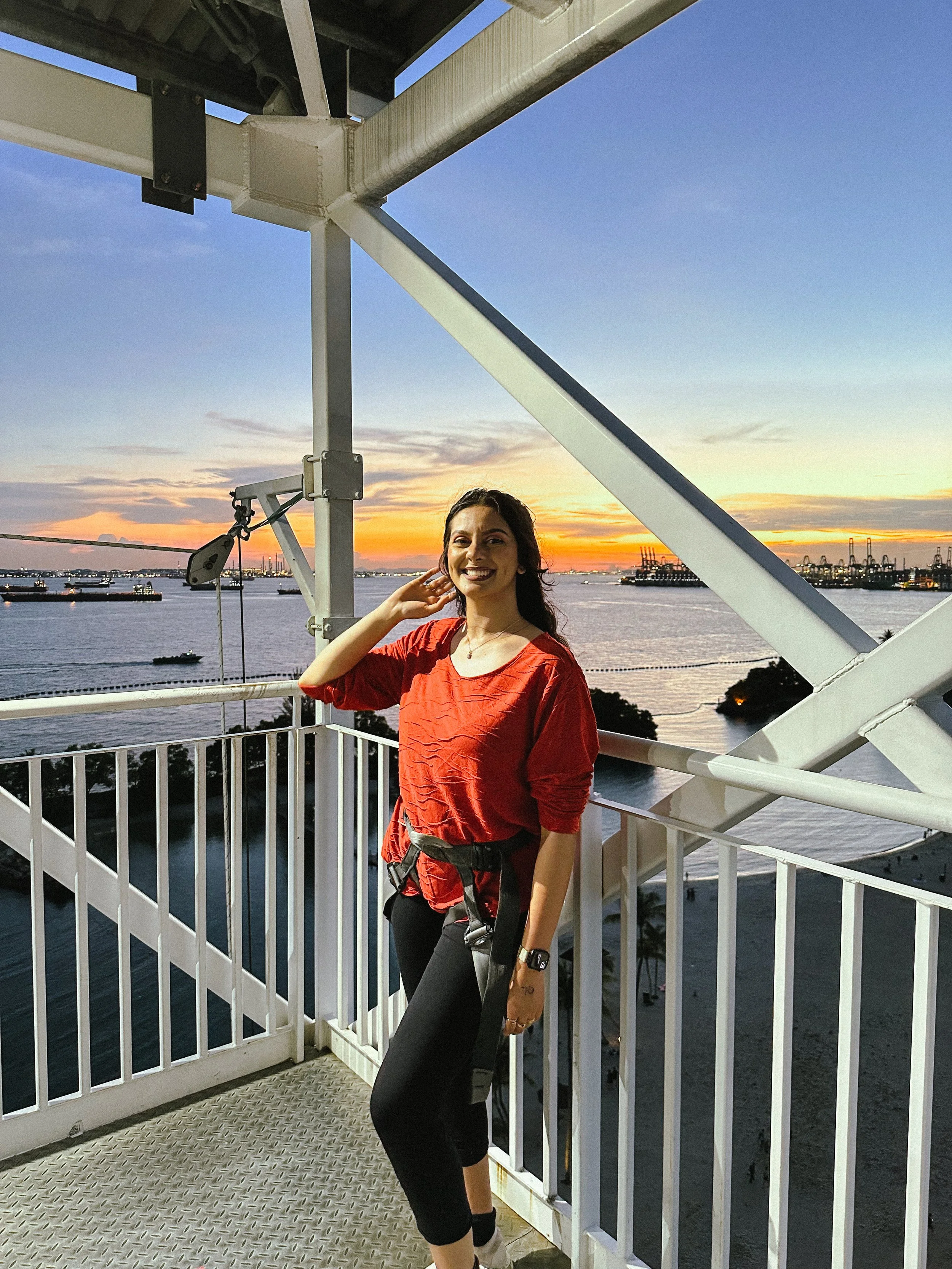 A woman standing on a ferris wheel at sunset over a body of water, smiling at the camera.