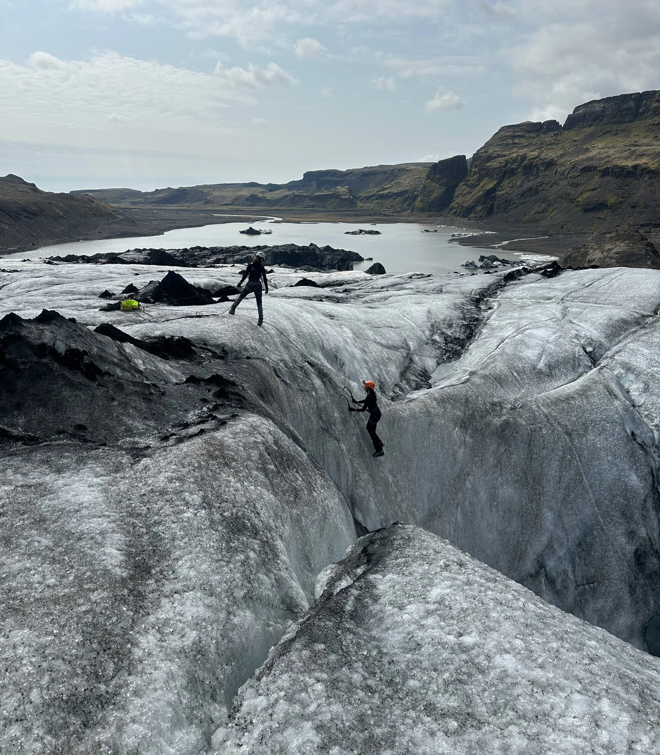 Two people are glacier climbing on a large ice sheet with a scenic river and mountainous landscape in the background.