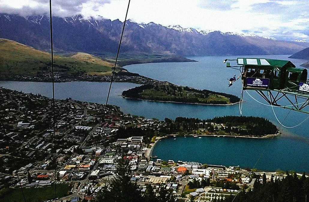 A cityscape viewed from a cable car, with lakes, green islands, and snow-capped mountains in the background.