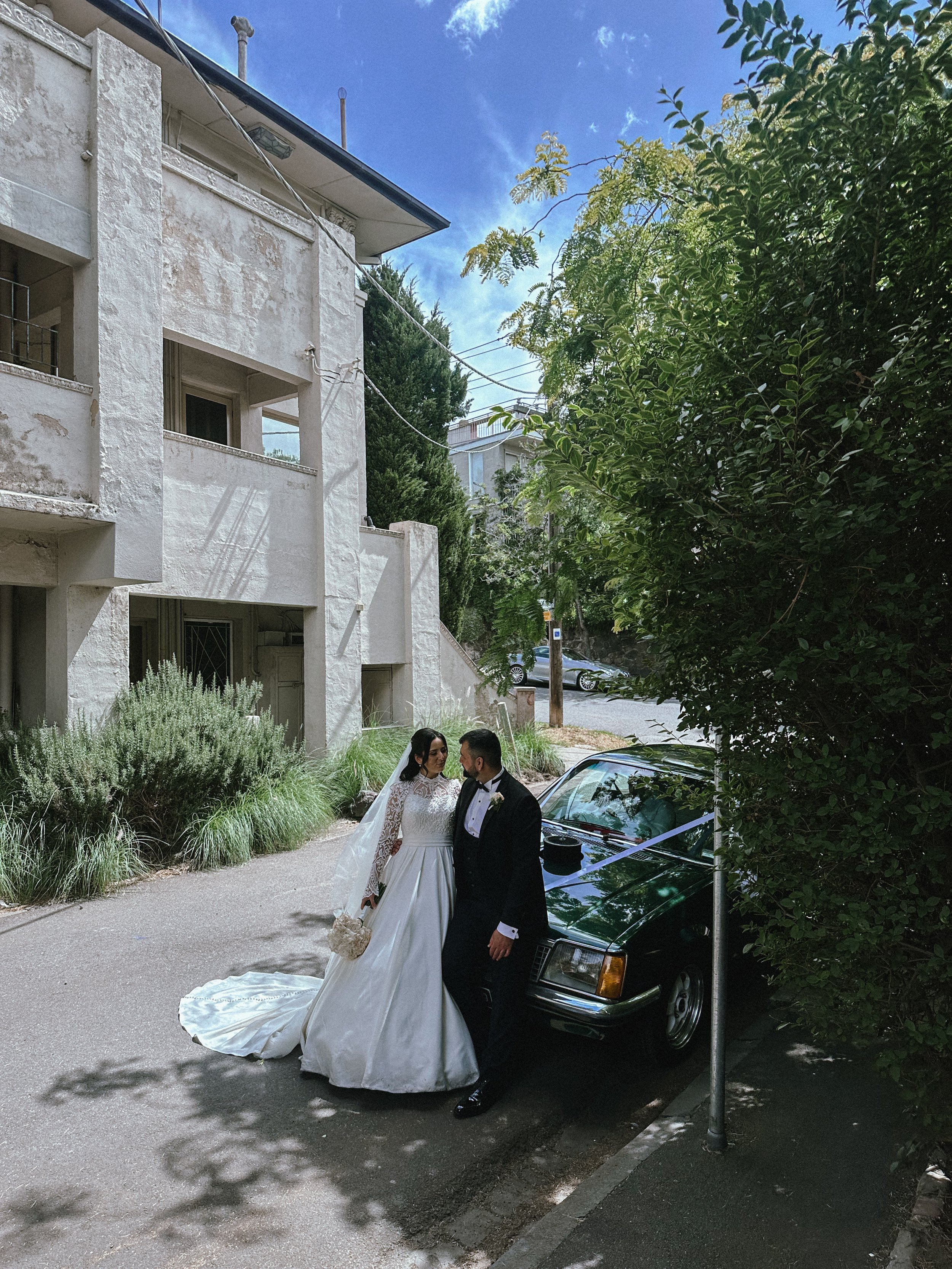 Bride and groom standing next to a black car on a street, with the bride in a white wedding dress and the groom in a tuxedo, surrounded by greenery and urban buildings.