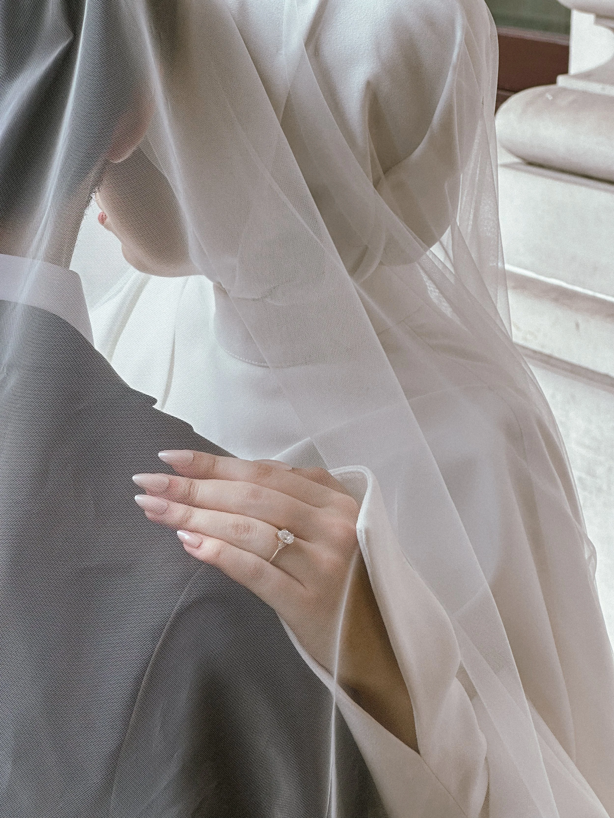 Close-up of a bride's hand with a diamond ring, gently resting on her groom's shoulder, behind a sheer white veil in a softly lit setting.