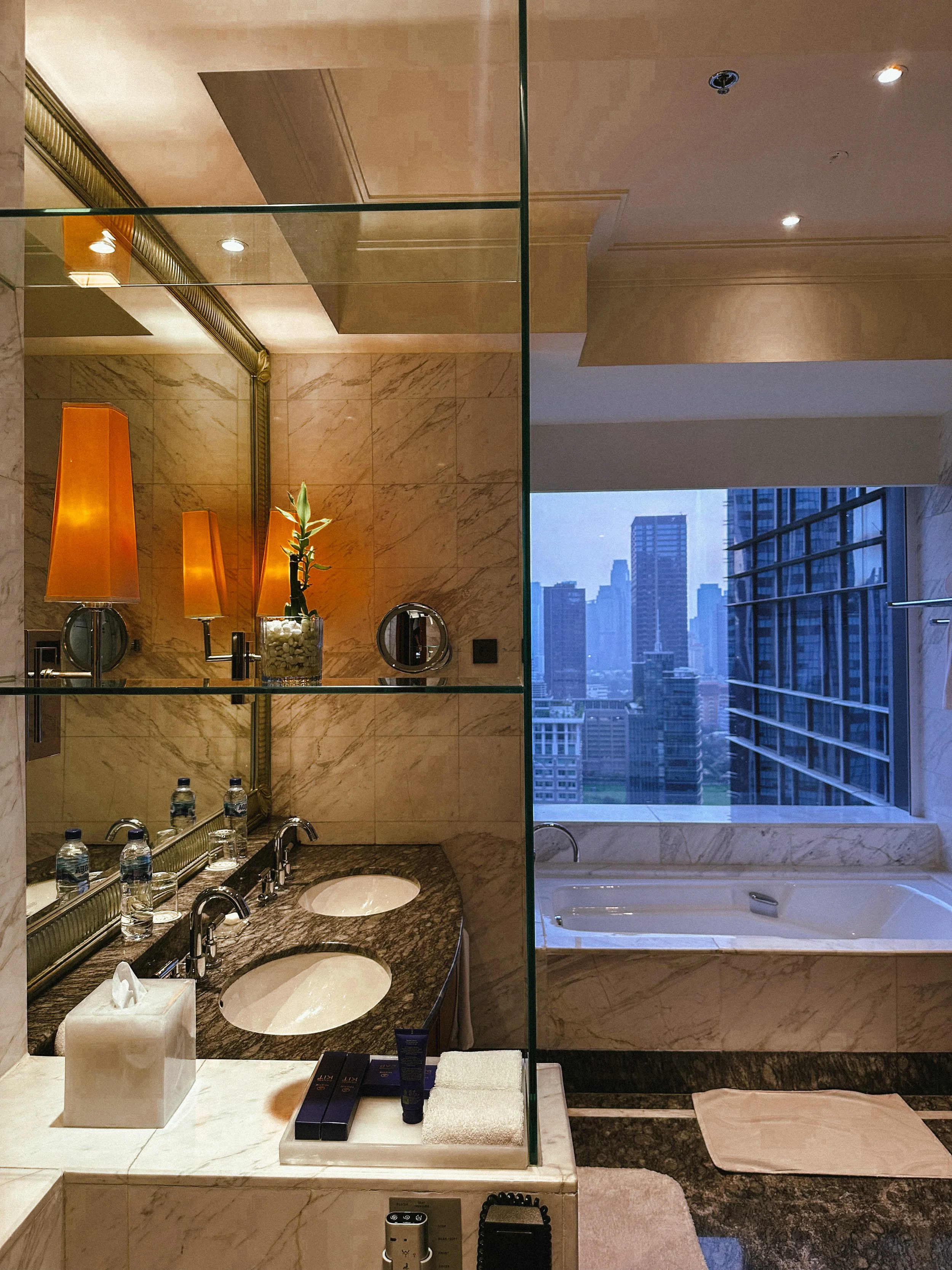 Luxury bathroom with marble countertops, dual sinks, a large window with a city view, two orange lamps, decorative plant, toiletries, tissues, and a bathtub.