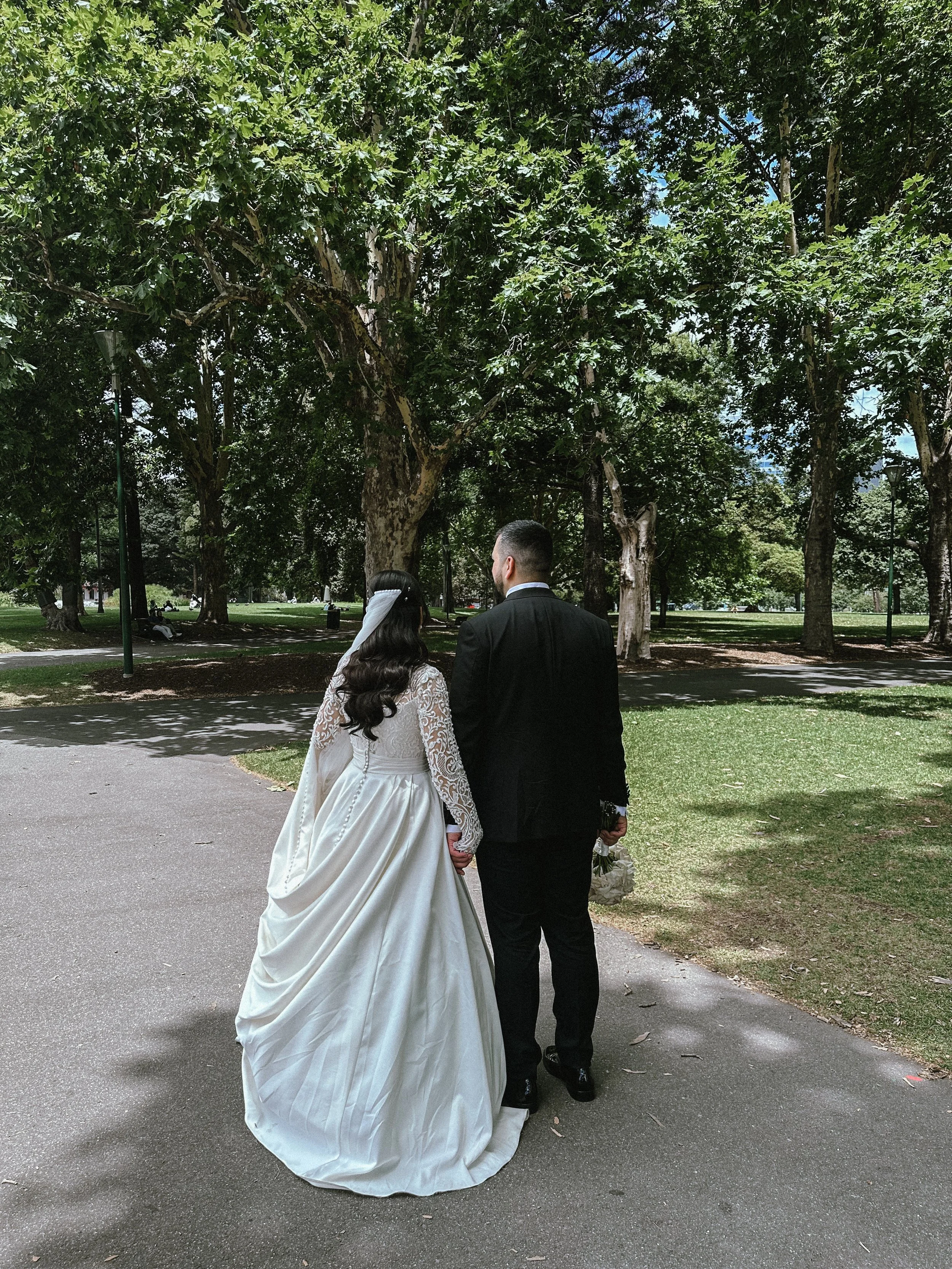 A bride and groom holding hands while walking in a park, with green trees and sunlight filtering through the leaves.