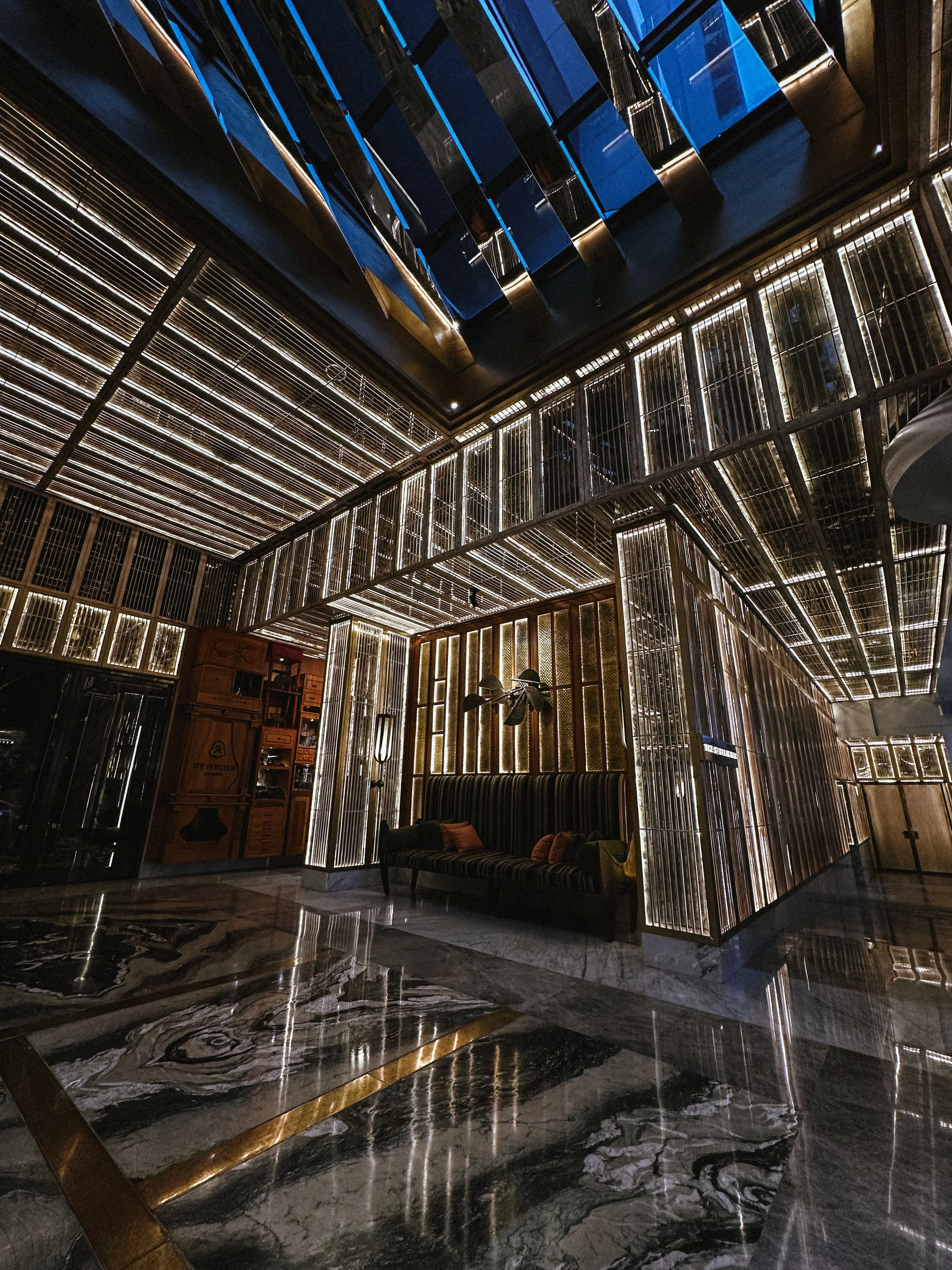 Interior of a modern building with illuminated wooden structure, a marble floor, a striped velvet sofa with pillows, and decorative lighting fixtures.