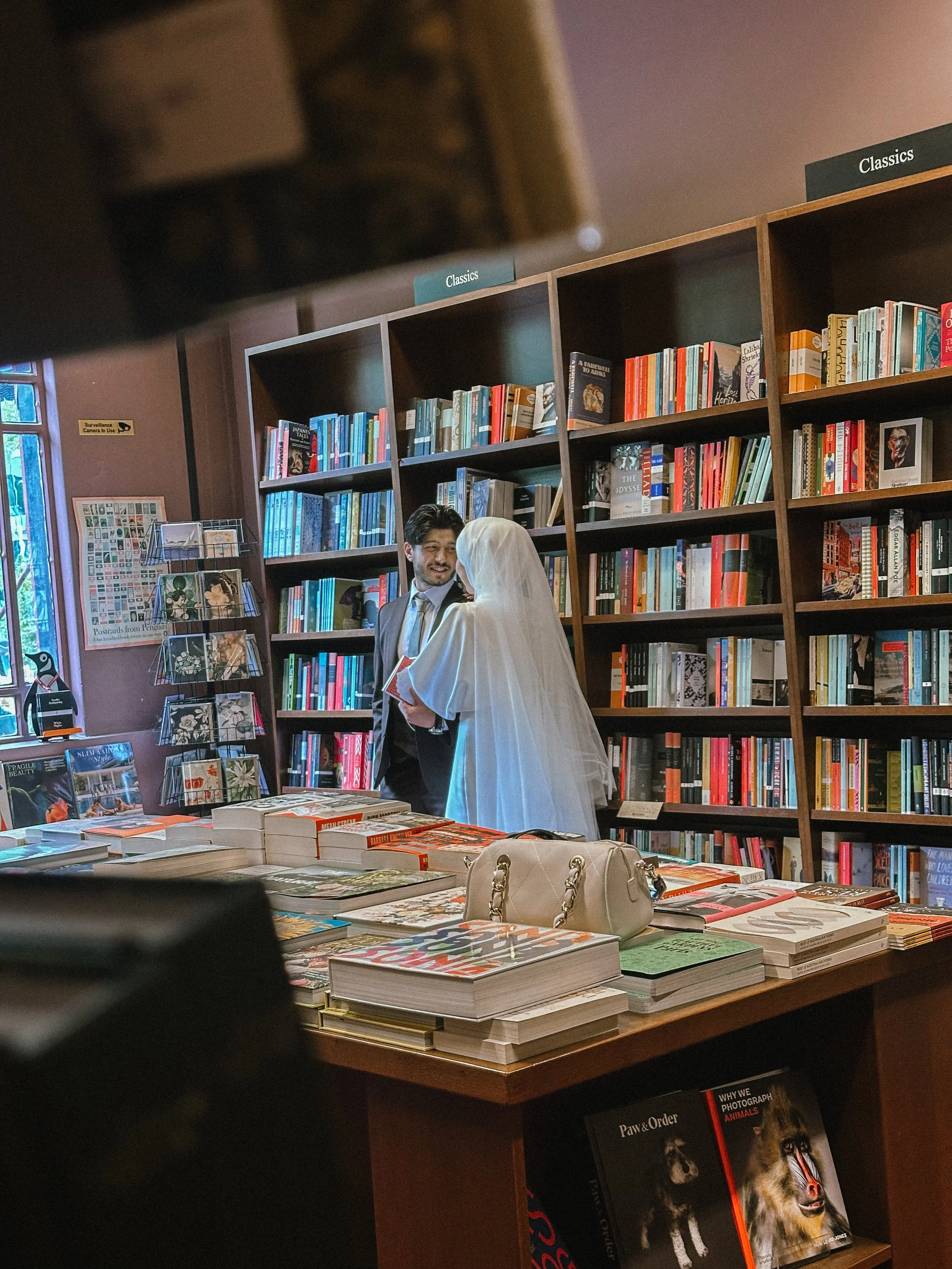 A couple in wedding attire browsing books in a bookstore, with bookshelves and a display table filled with books in the background.