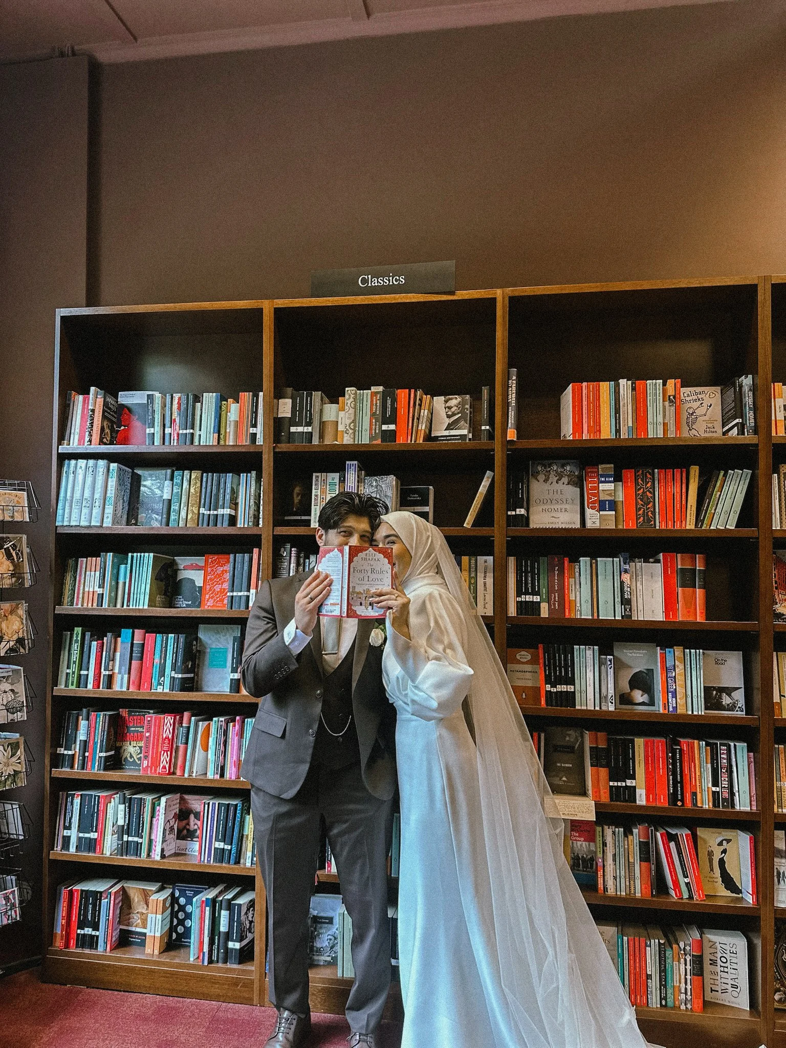 A man in a suit and a woman in a wedding dress reading a book together in front of a bookshelf labeled 'Classics' in a bookstore.