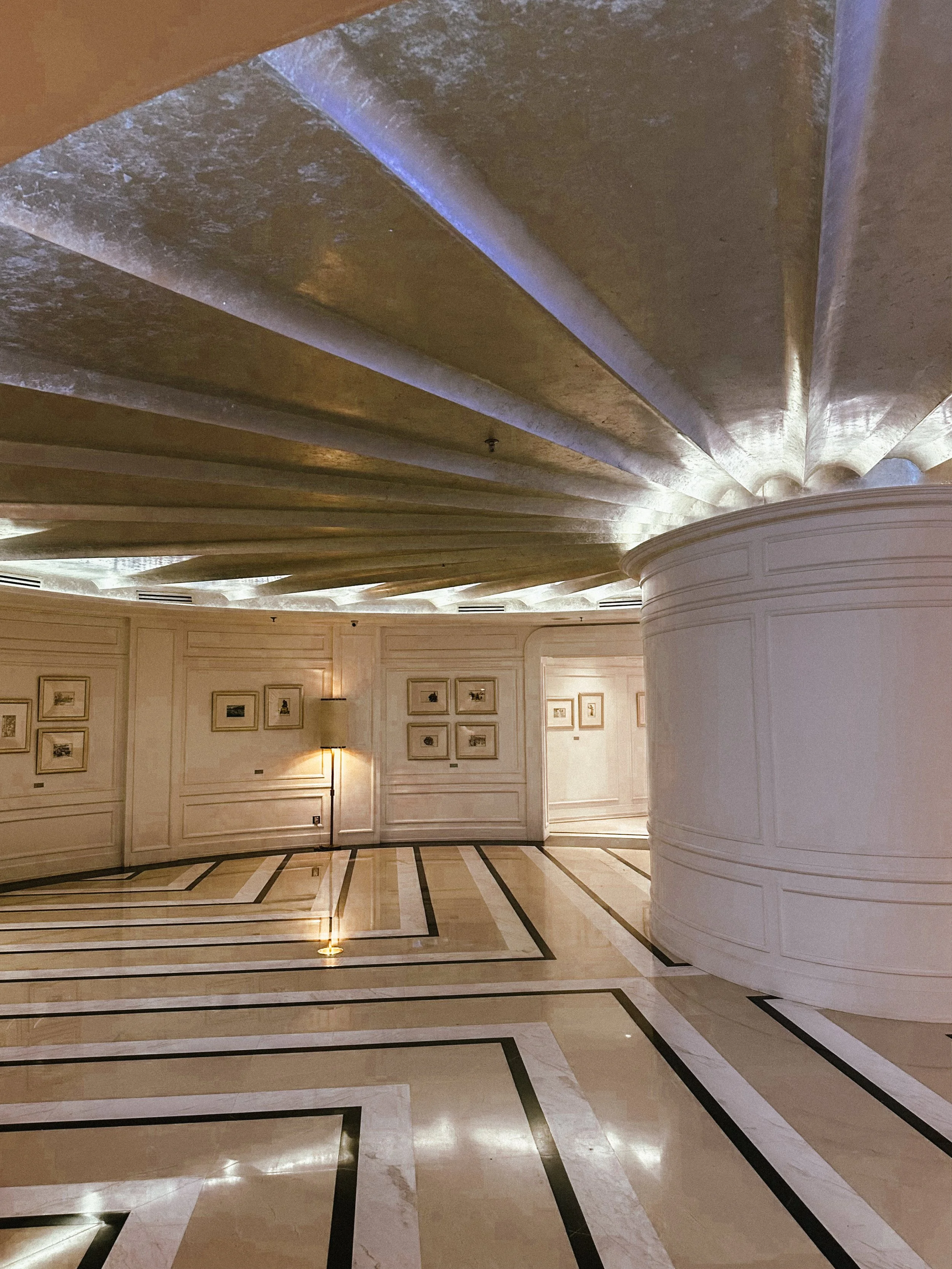 Elegant hotel lobby with marble flooring, framed artwork on cream-colored walls, a rounded white reception desk, and a decorative gold ceiling.