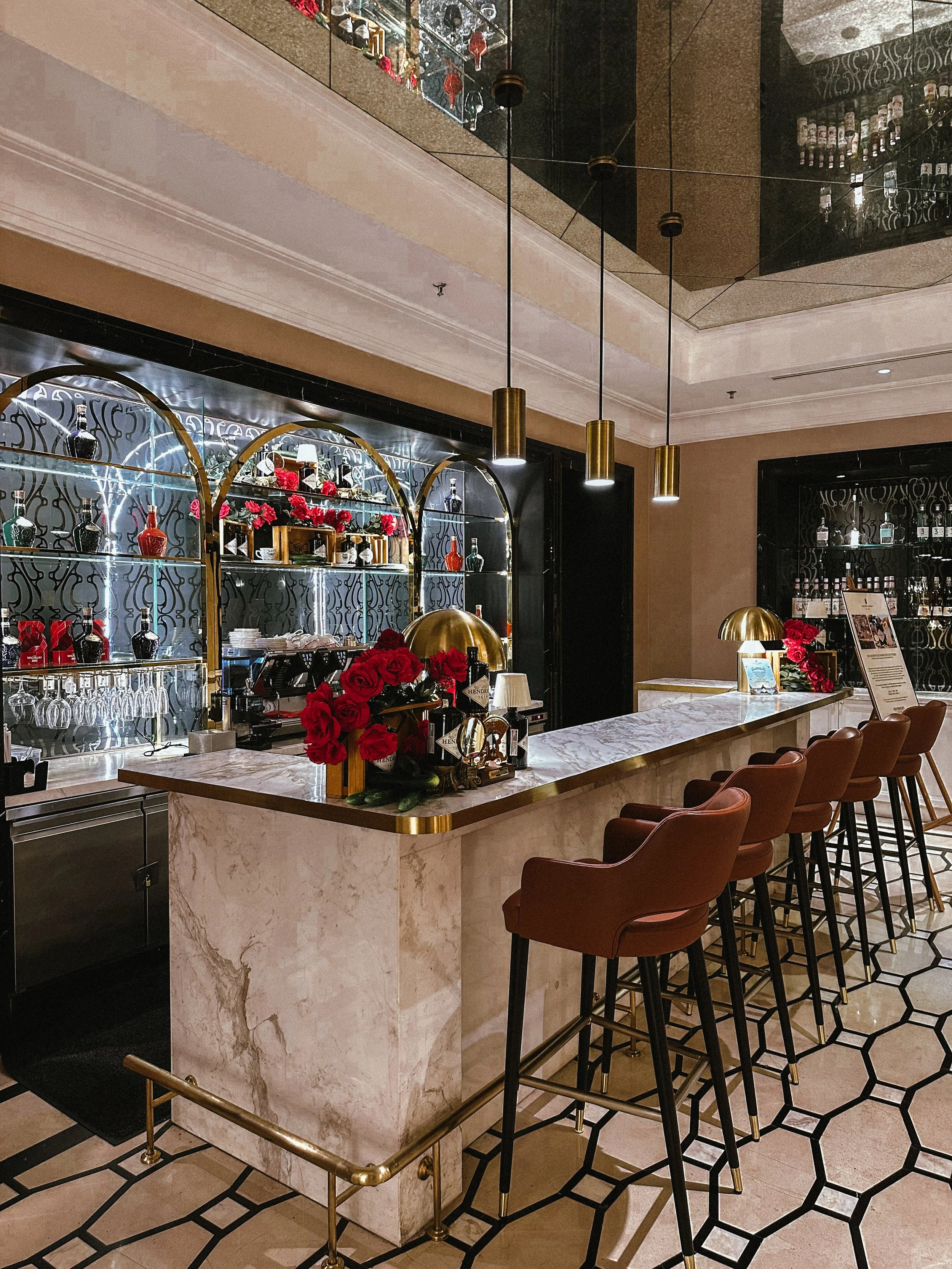 Elegant hotel bar with a marble counter, bar stools, gold accents, and shelves with liquor bottles and glasses.