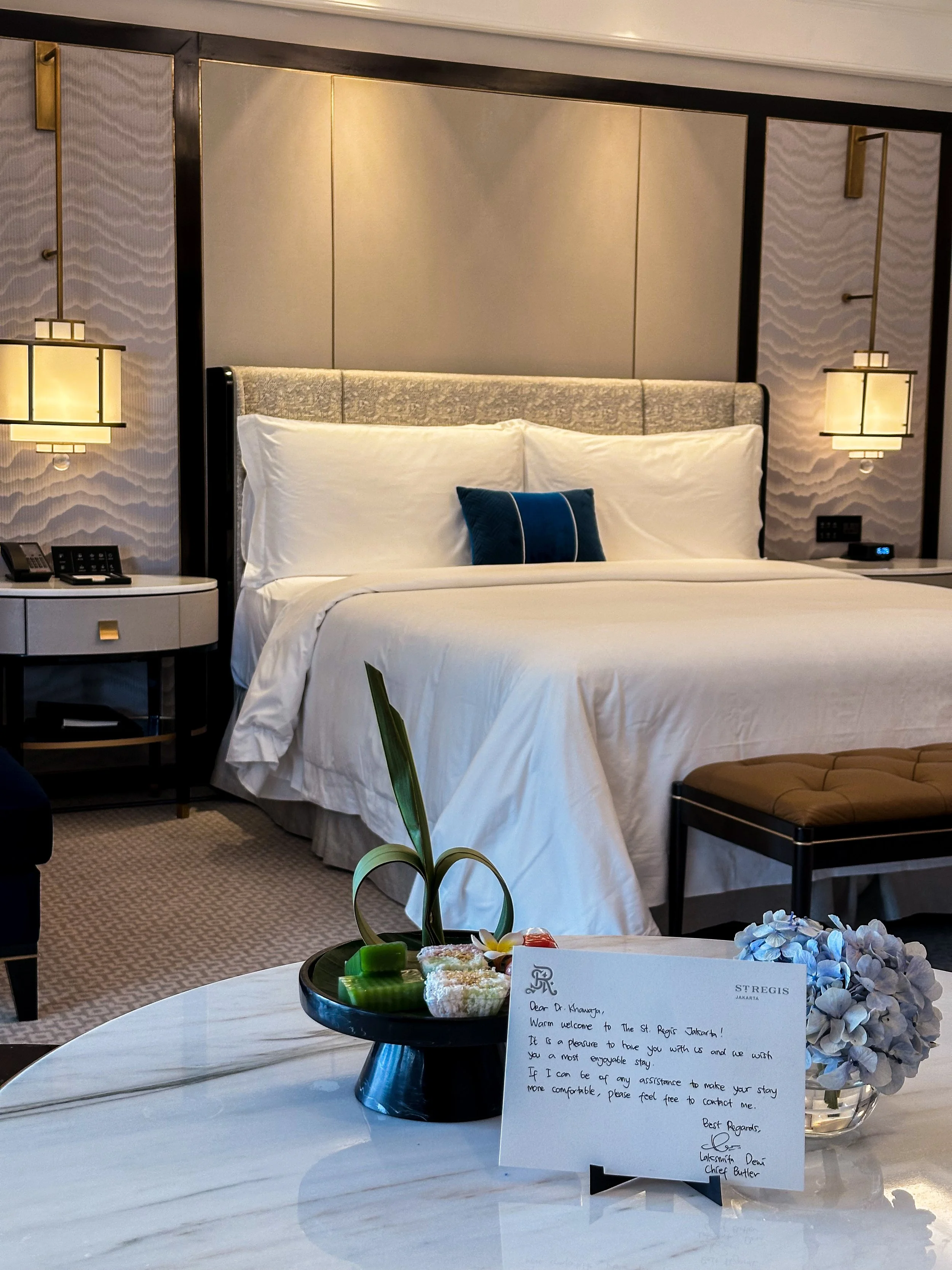 A hotel room with a made bed, bedside lamps, and a table with a welcome note, a flower arrangement, and snacks.